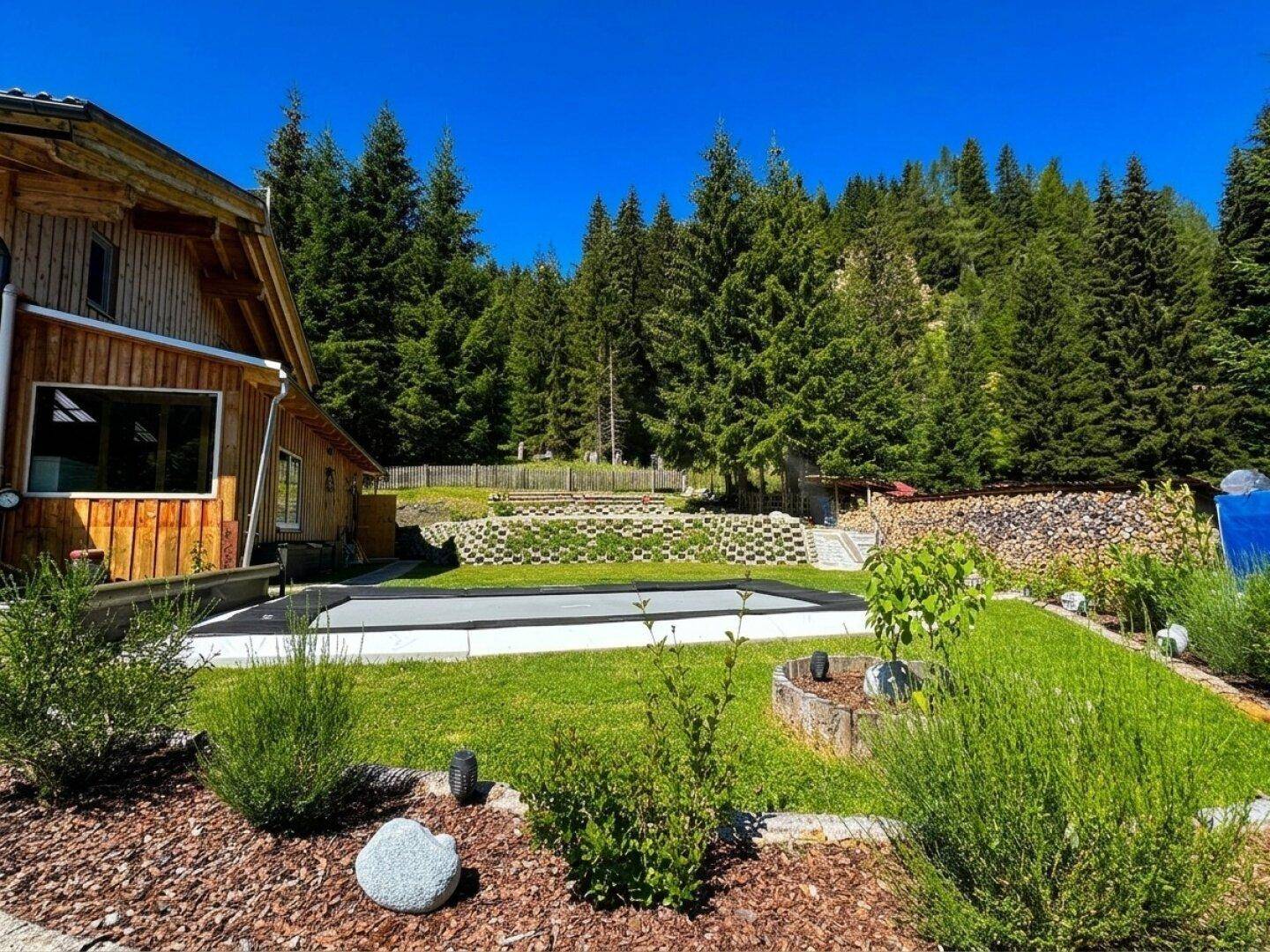 Grüner Gartenbereich mit eingelassenem Trampolin oder Pool und Holzstapel im Hintergrund.