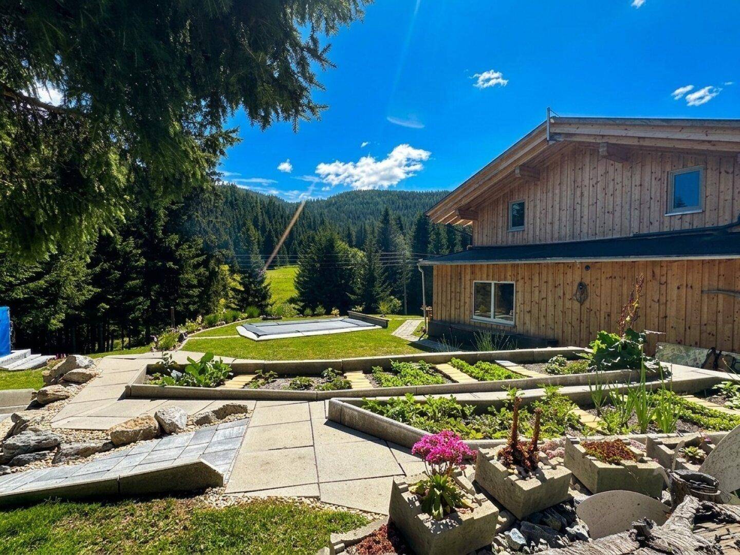Terrassierter Garten mit vielfältigen Pflanzen, gepflasterten Wegen und Blick auf das Haus und die Berge.