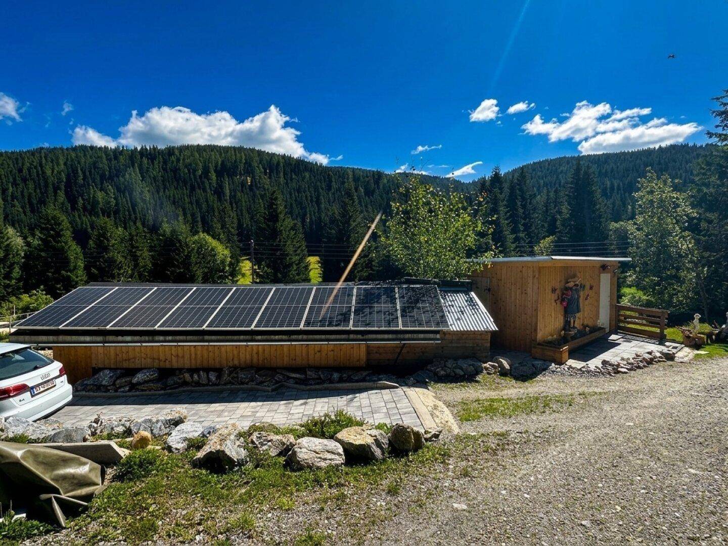 Holzgebäude mit Photovoltaikanlage auf dem Dach, umgeben von grünen Wäldern und Bergen unter blauem Himmel.