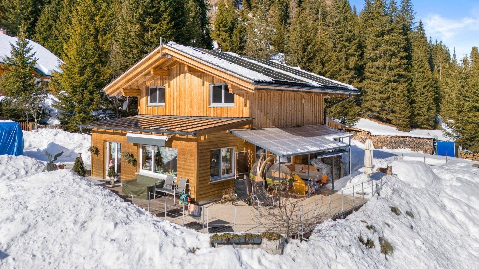 Holzhaus mit überdachter Terrasse und Sitzgelegenheiten, umgeben von Schnee und Wald in den Bergen.