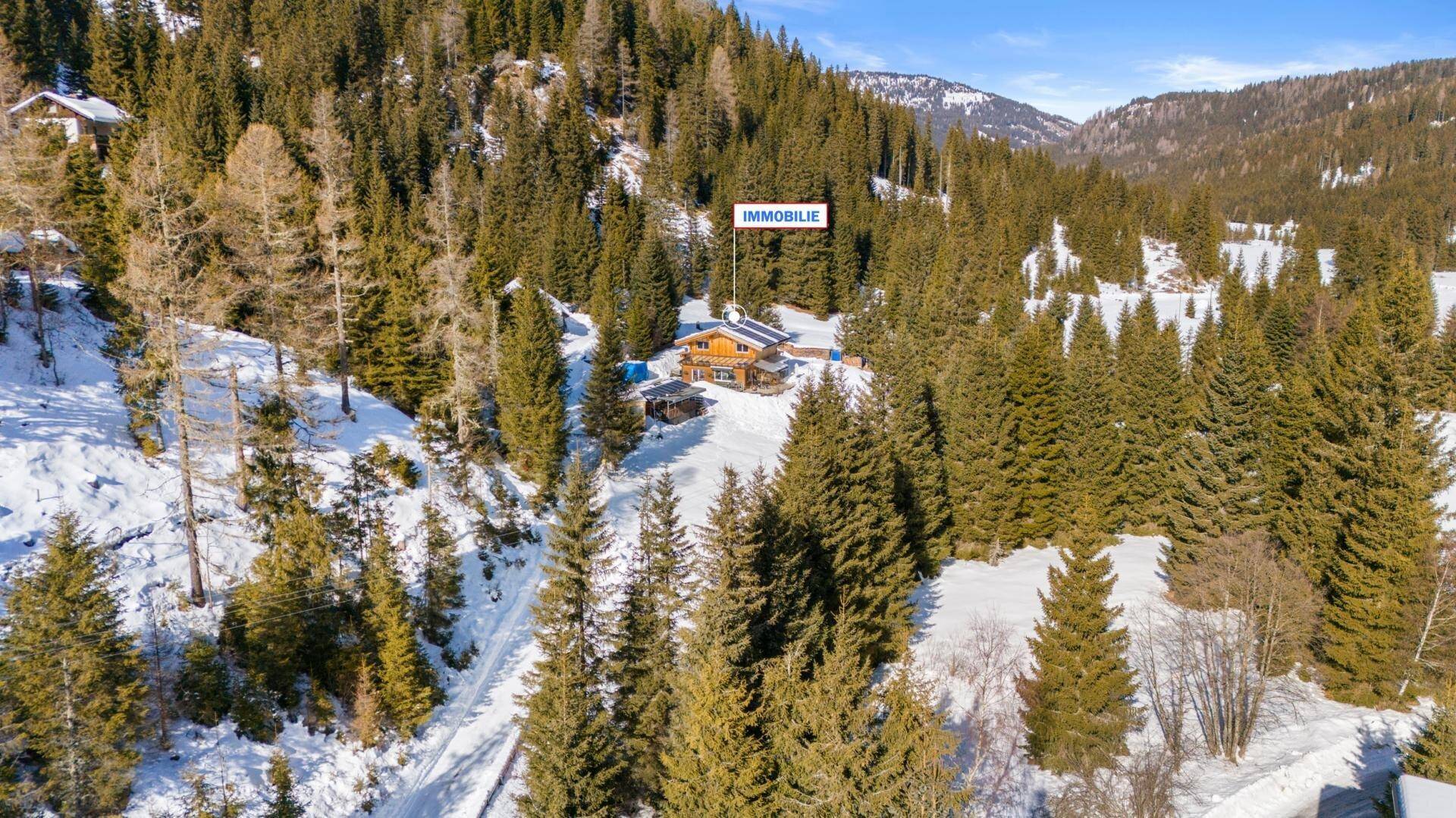 Luftaufnahme eines Holzhauses in einer verschneiten Berglandschaft, umgeben von dichten Nadelwäldern.