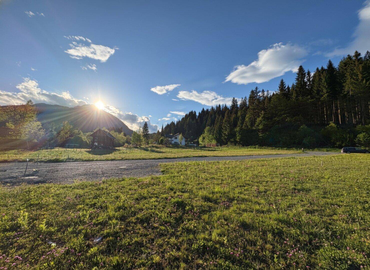 Sonnenuntergang über einer weiten Wiese mit Bergen und Wald im Hintergrund, einige Häuser sind sichtbar.