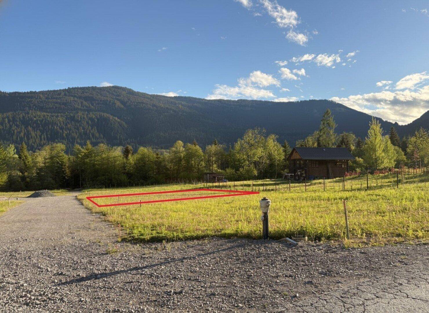 Eine grüne Wiese mit einer roten Grundstücksmarkierung, dahinter Berge und ein traditionelles Holzhaus.
