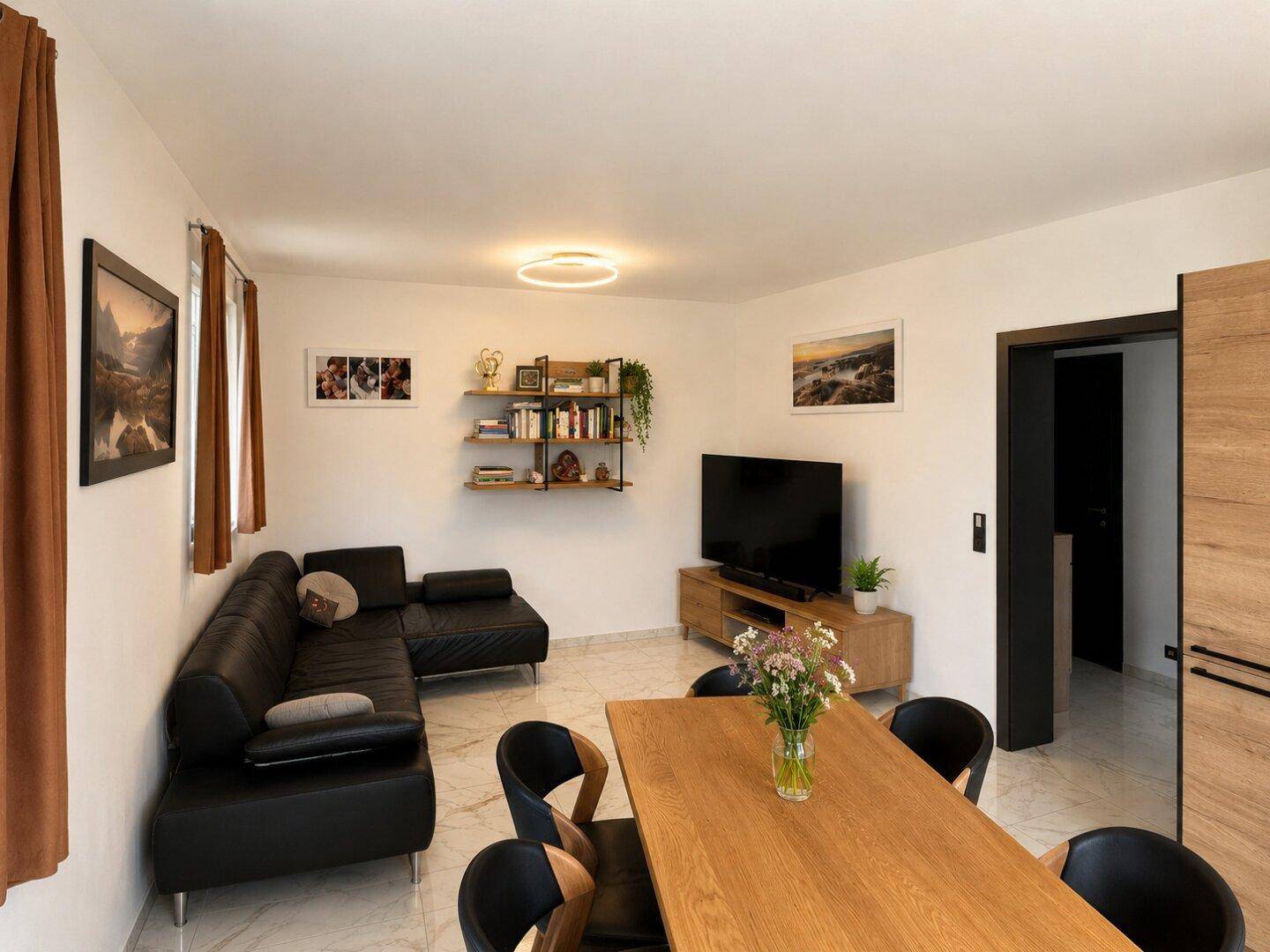 Gemütlicher Wohn- und Essbereich mit schwarzem Sofa, Holztisch und modernen Fliesenboden.