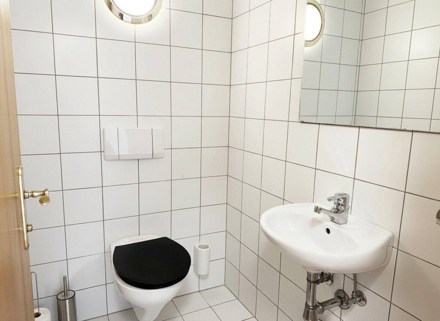 Kleines WC mit weißen Fliesen, Toilette, Einzelwaschbecken und zwei runden Fenstern.
