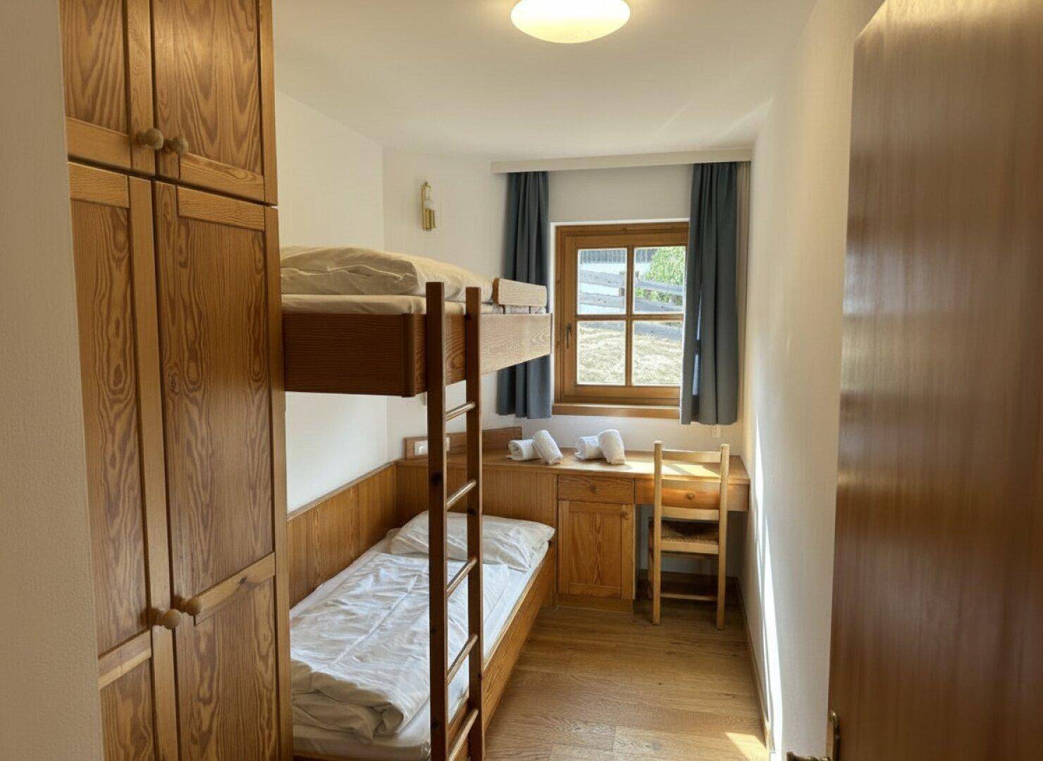 Kinderzimmer mit Etagenbett, Schreibtisch und Holzschränken, hell durch ein Fenster.
