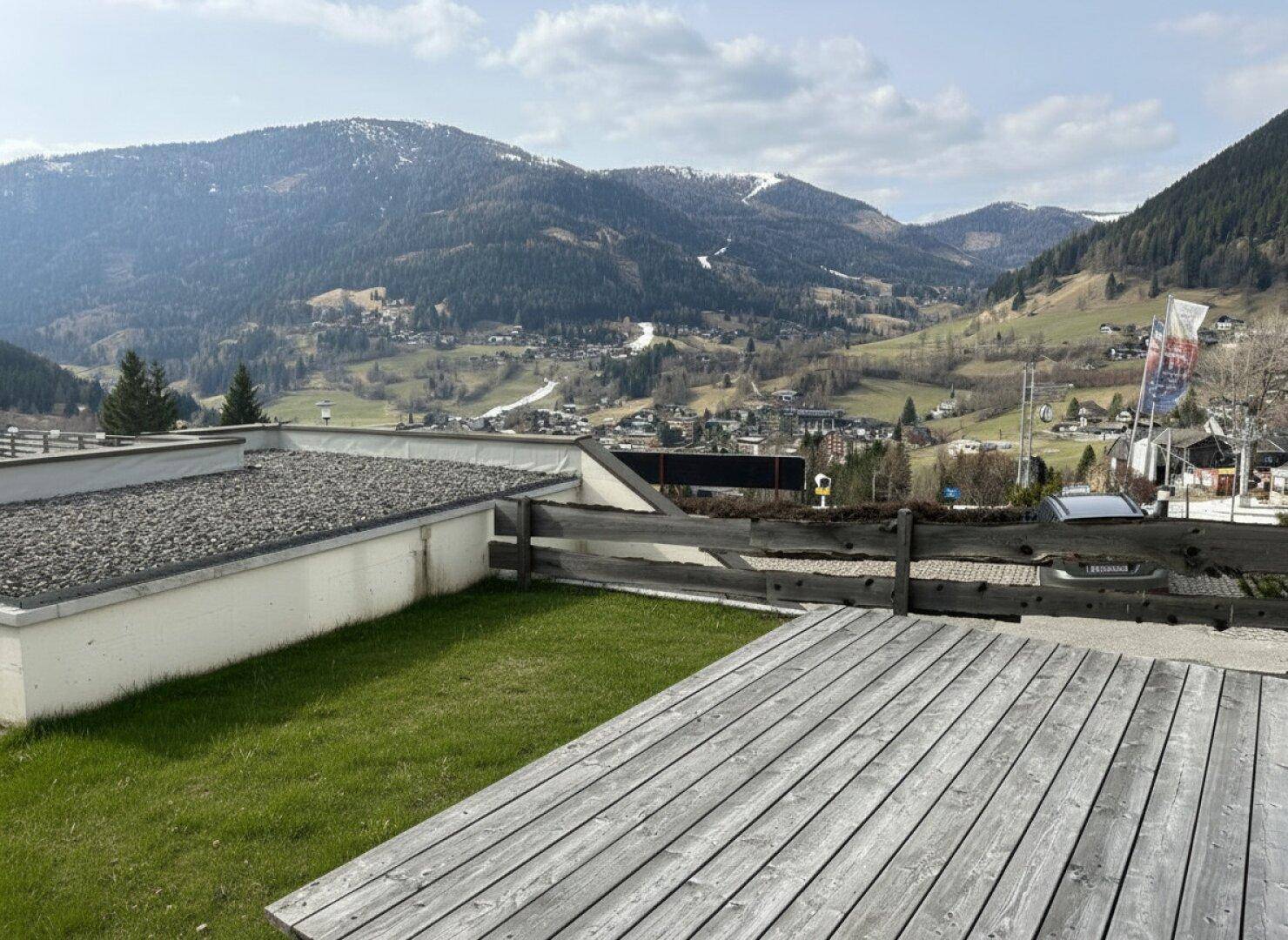 Sonniger Gartenbereich mit Holzterrasse und Rasenfläche, umgeben von Bergen.