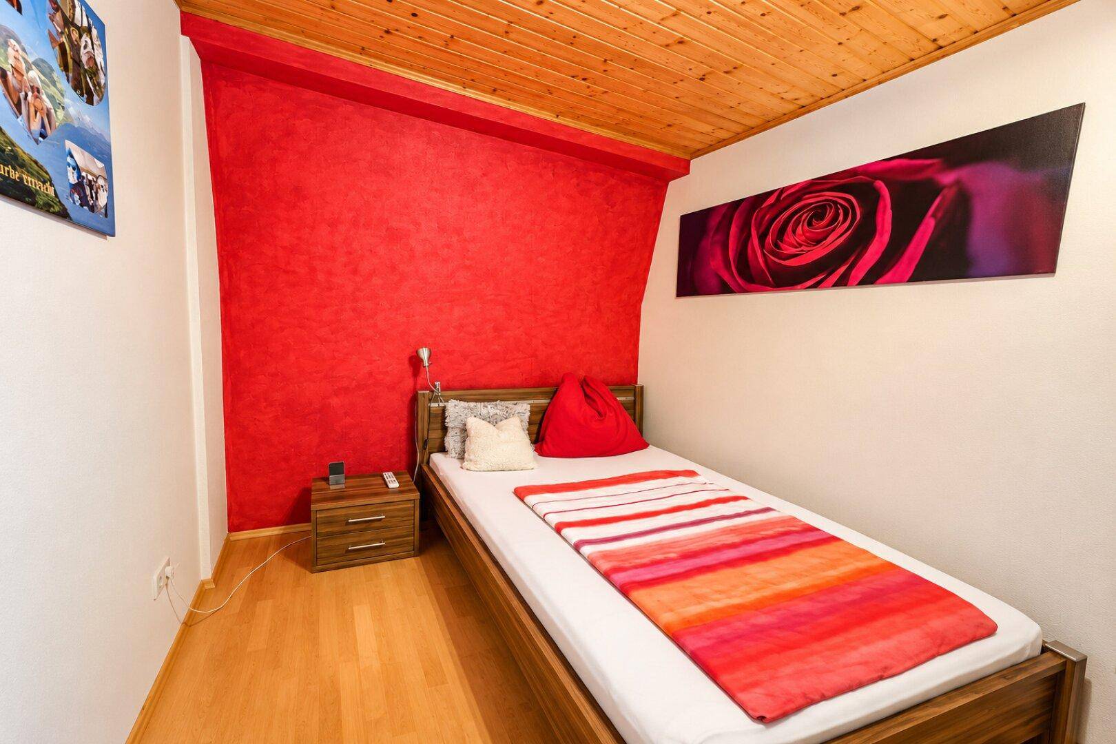 Kleines Schlafzimmer mit Einzelbett, roter Akzentwand und Holzdecke.