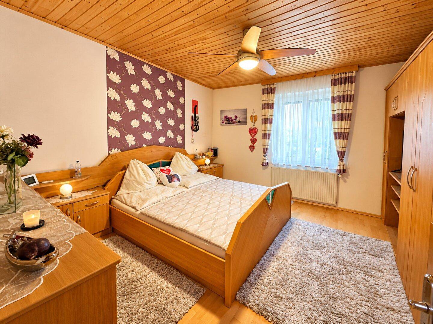 Schlafzimmer mit Holztäfelung an der Decke, Doppelbett und floraler Tapete.