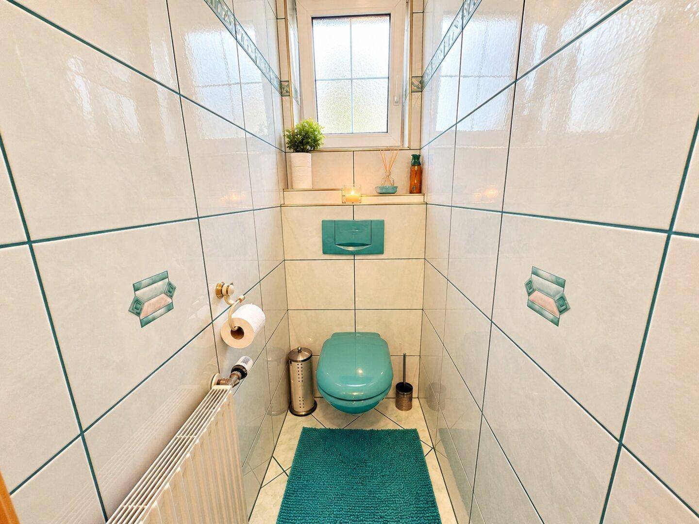 Separate Toilette mit hellen Fliesen, Fenster und farbiger WC-Schüssel.