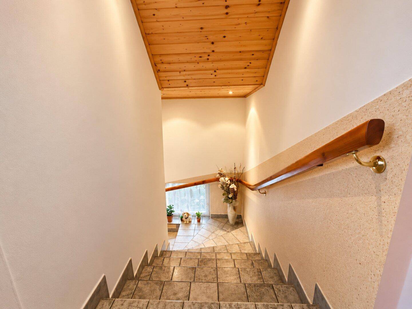 Treppenhaus mit Holzgeländer, Fliesenboden und Holzdecke.