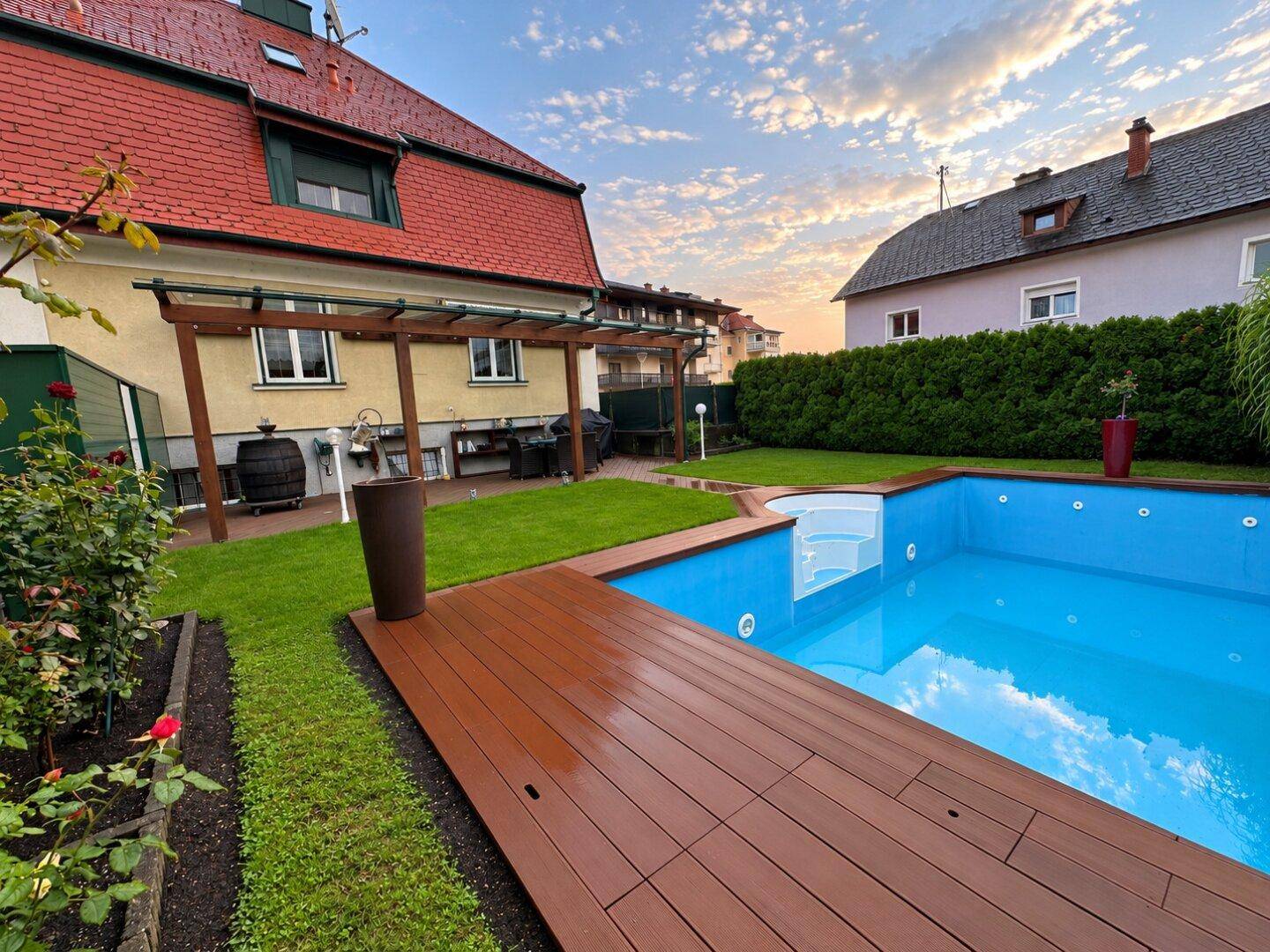 Gepflegter Gartenbereich mit Swimmingpool, Holzterrasse und Pergola unter einem schönen Abendhimmel.
