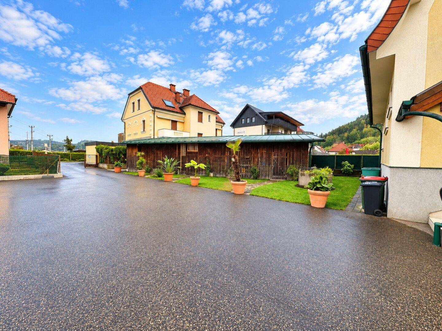 Zufahrt mit gepflastertem Bereich, einem Holzschuppen und einem Wohnhaus im Hintergrund mit grüner Aussicht.