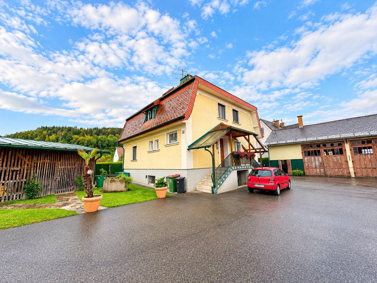 Gelbes Wohnhaus mit rotem Dach, gepflasterter Zufahrt und einem roten Auto vor einem Holzschuppen.