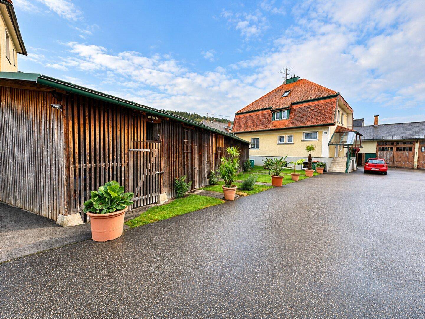 Holzschuppen mit gepflastertem Vorplatz und Topfpflanzen, dahinter ein gelbes Wohnhaus mit rotem Dach.