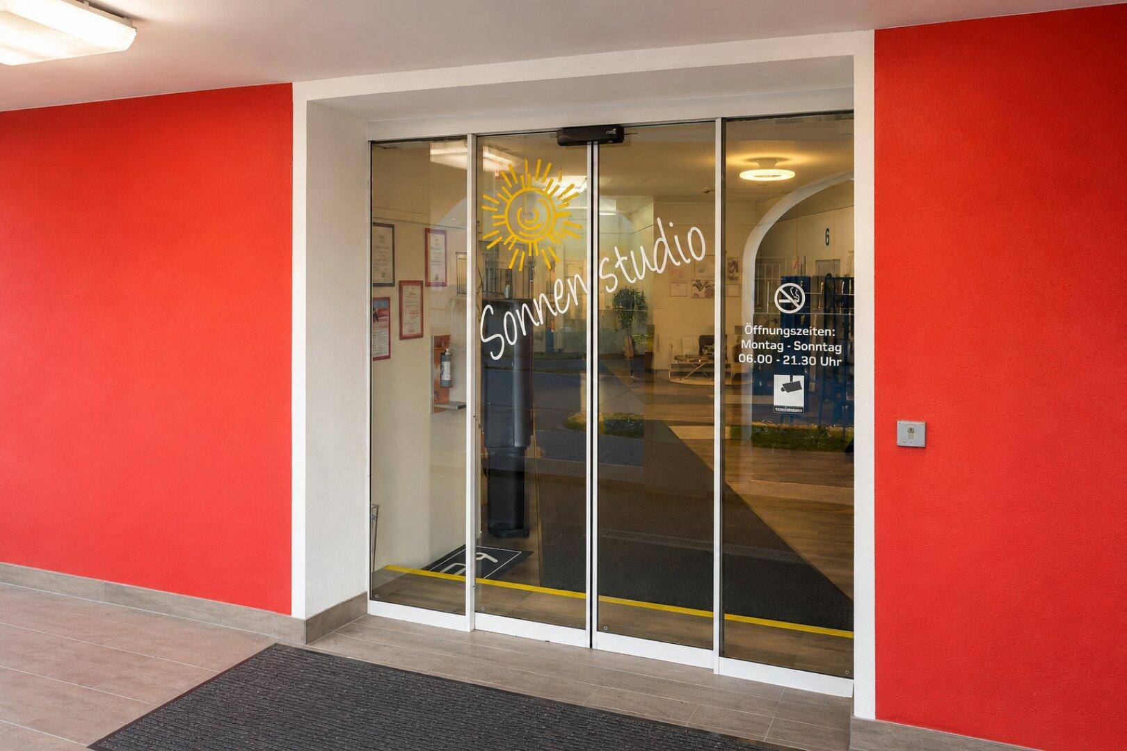 Eingangsbereich eines Sonnenstudios mit Glastüren, roten Wänden und Öffnungszeiten-Aufklebern.
