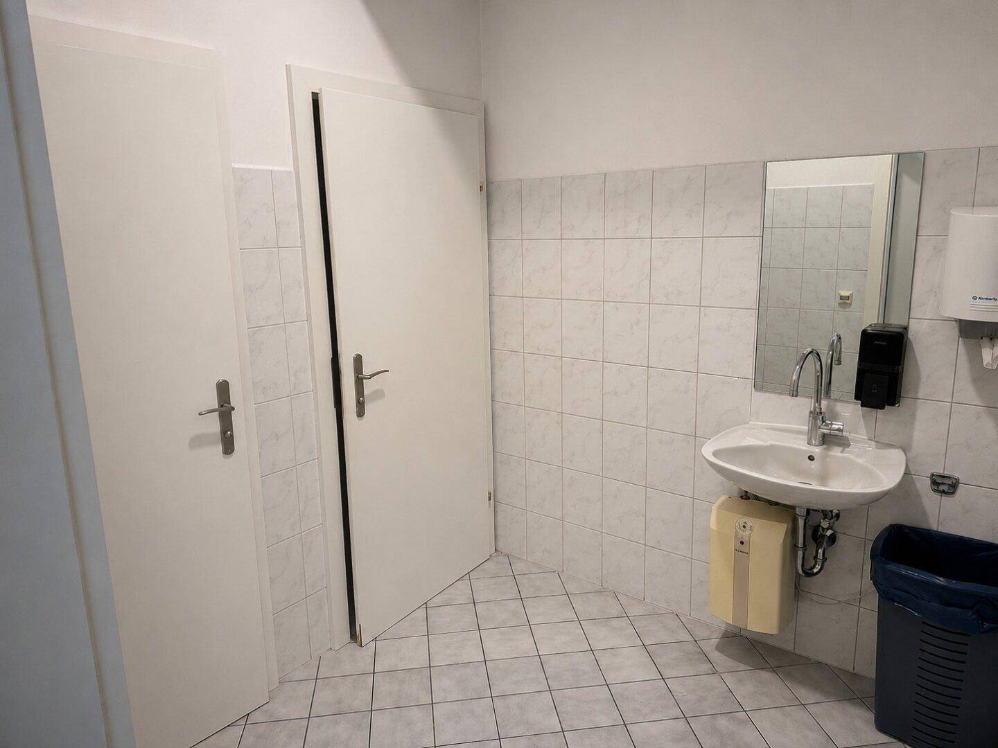 Einfache Toilette mit weißen Fliesen, Waschbecken, Spiegel und zwei Türen.