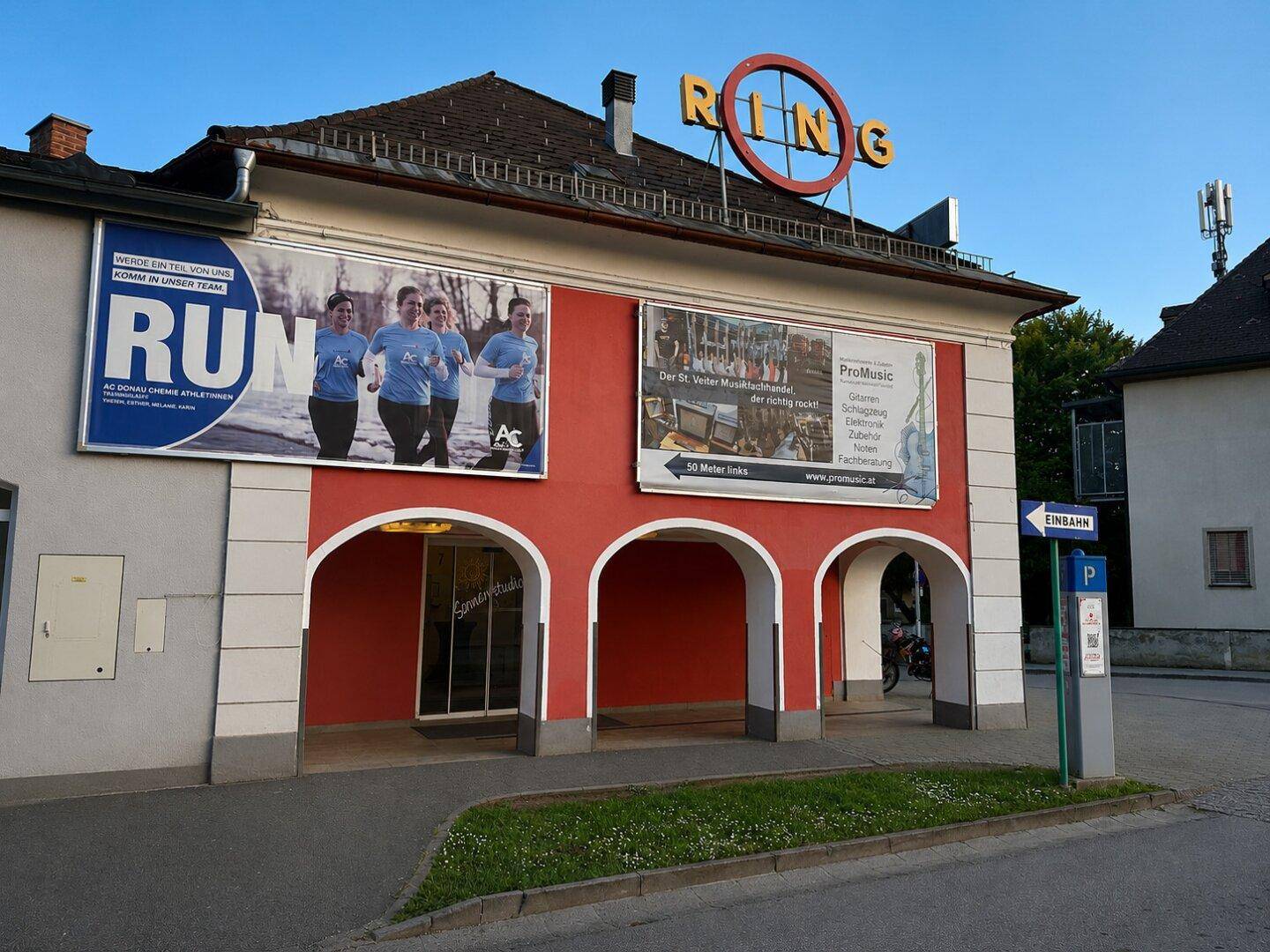 Außenansicht eines Gebäudes mit roten Arkaden, Werbebannern und einem "RING"-Schild.