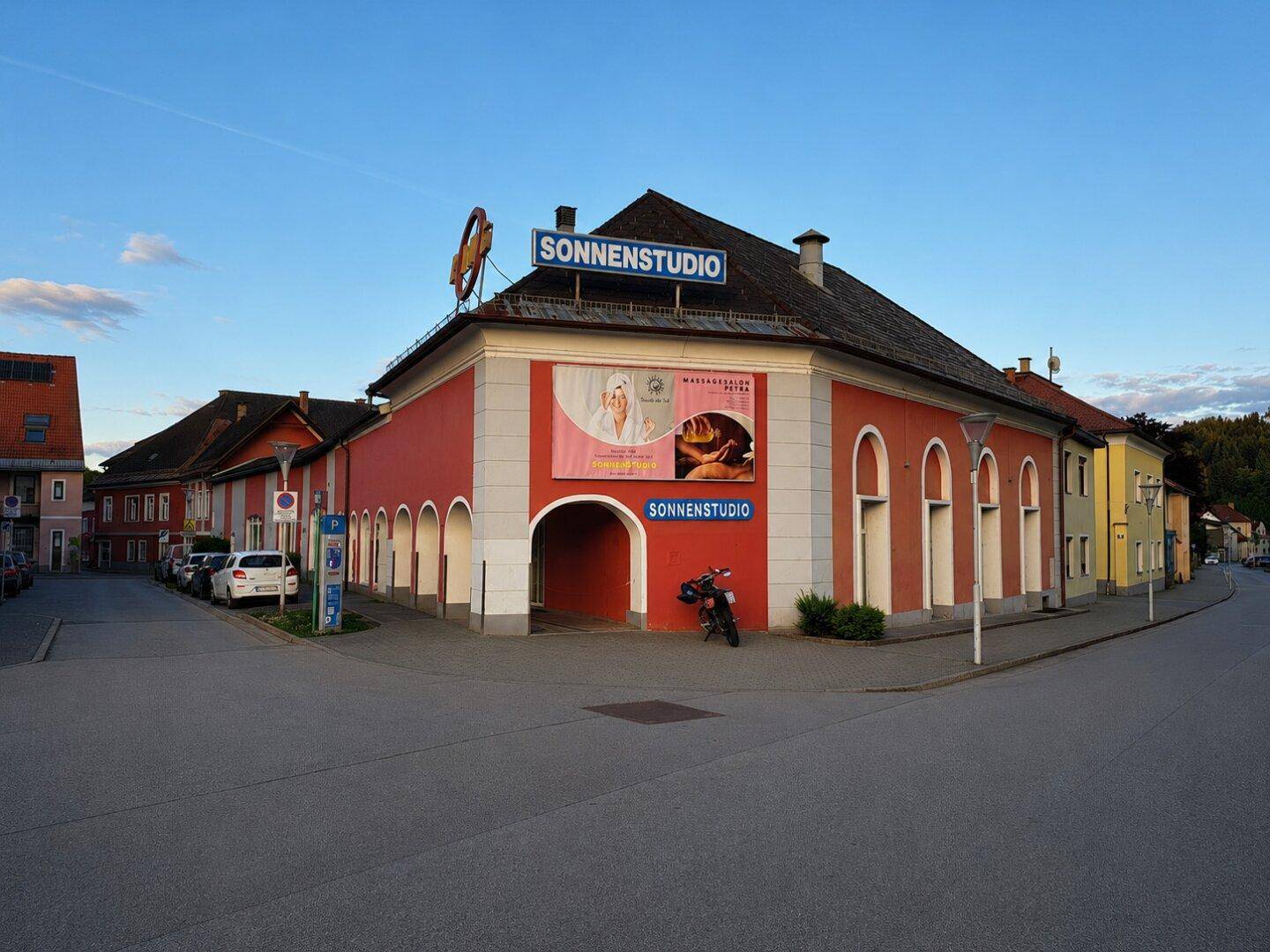 Weitläufige Außenansicht eines Gebäudes mit roten Arkaden, Sonnenstudio-Schild und umliegender Straße.