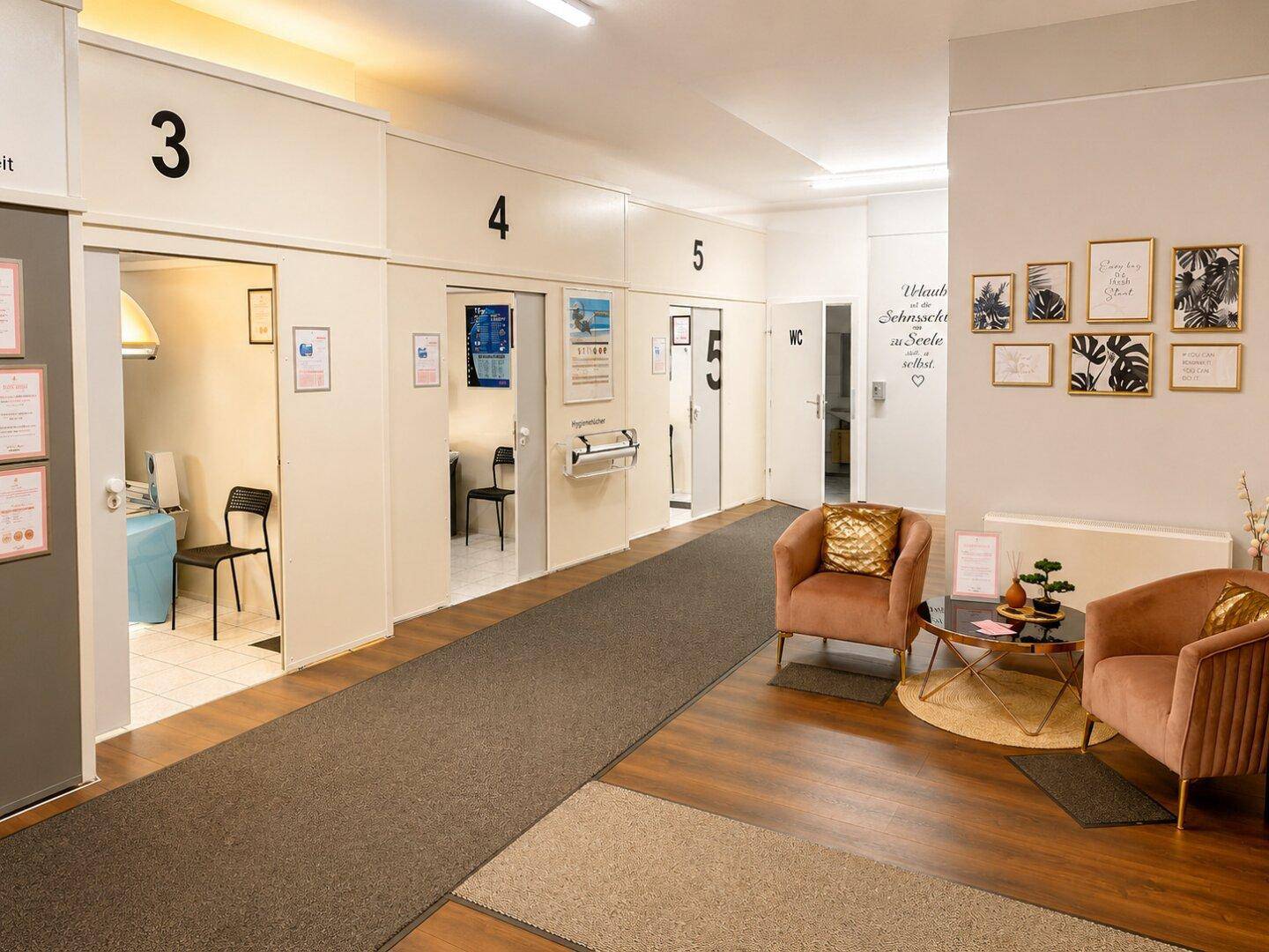 Heller Flur mit nummerierten Kabinen, einer Sitzecke und gerahmten Bildern an der Wand.
