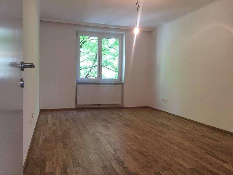Leeres, helles Zimmer mit Parkettboden, weißen Wänden und einem Fenster mit grüner Aussicht.