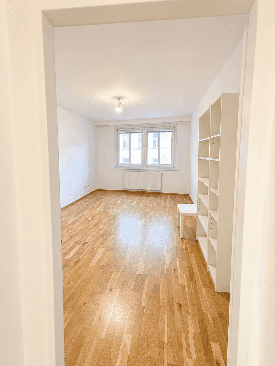 Helles, leeres Zimmer mit Holzparkettboden, weißen Wänden und einem großen Fenster.