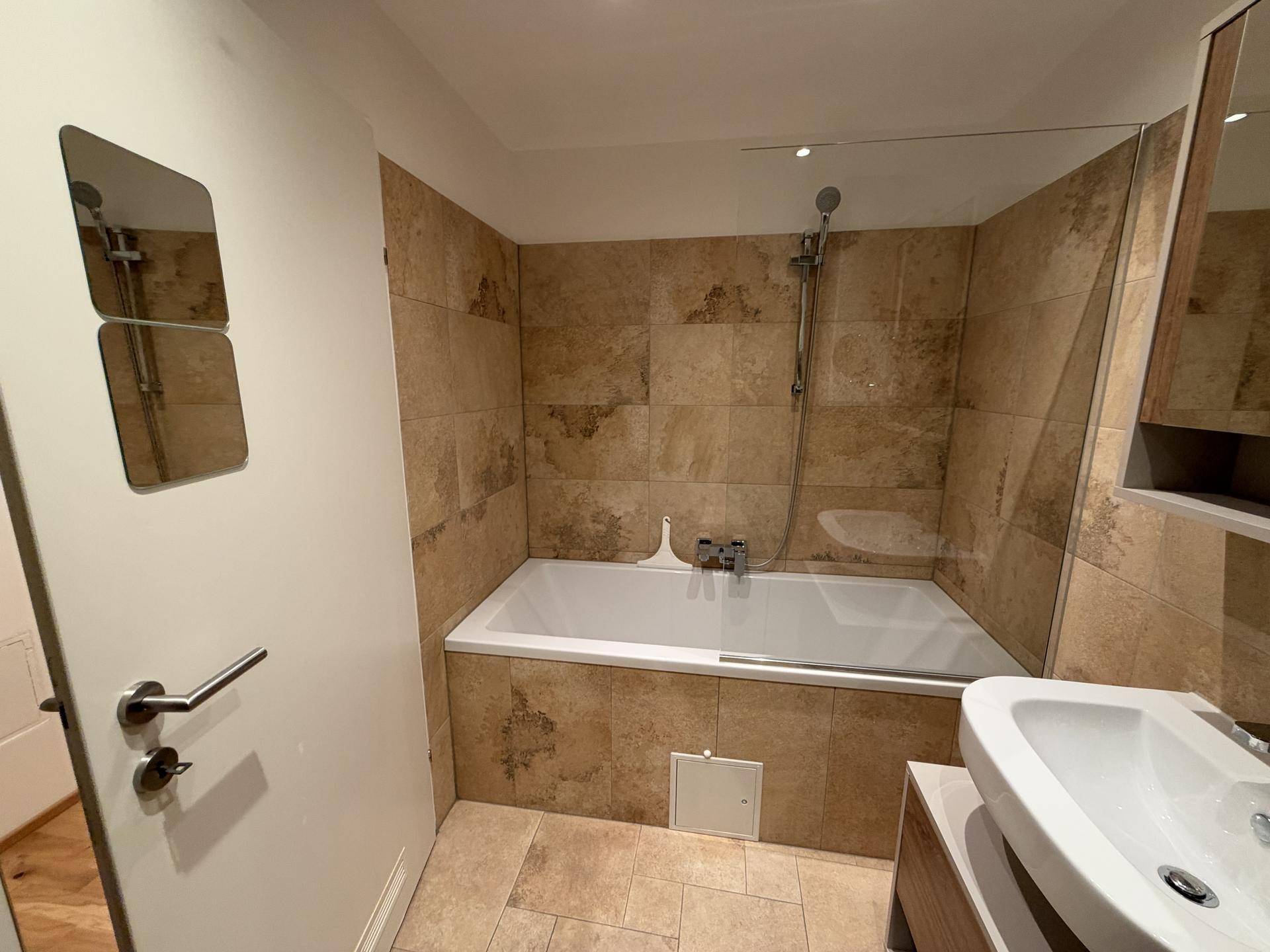 Gefliestes Badezimmer mit Badewanne und Duschvorrichtung, ausgestattet mit einer Glasabtrennung.
