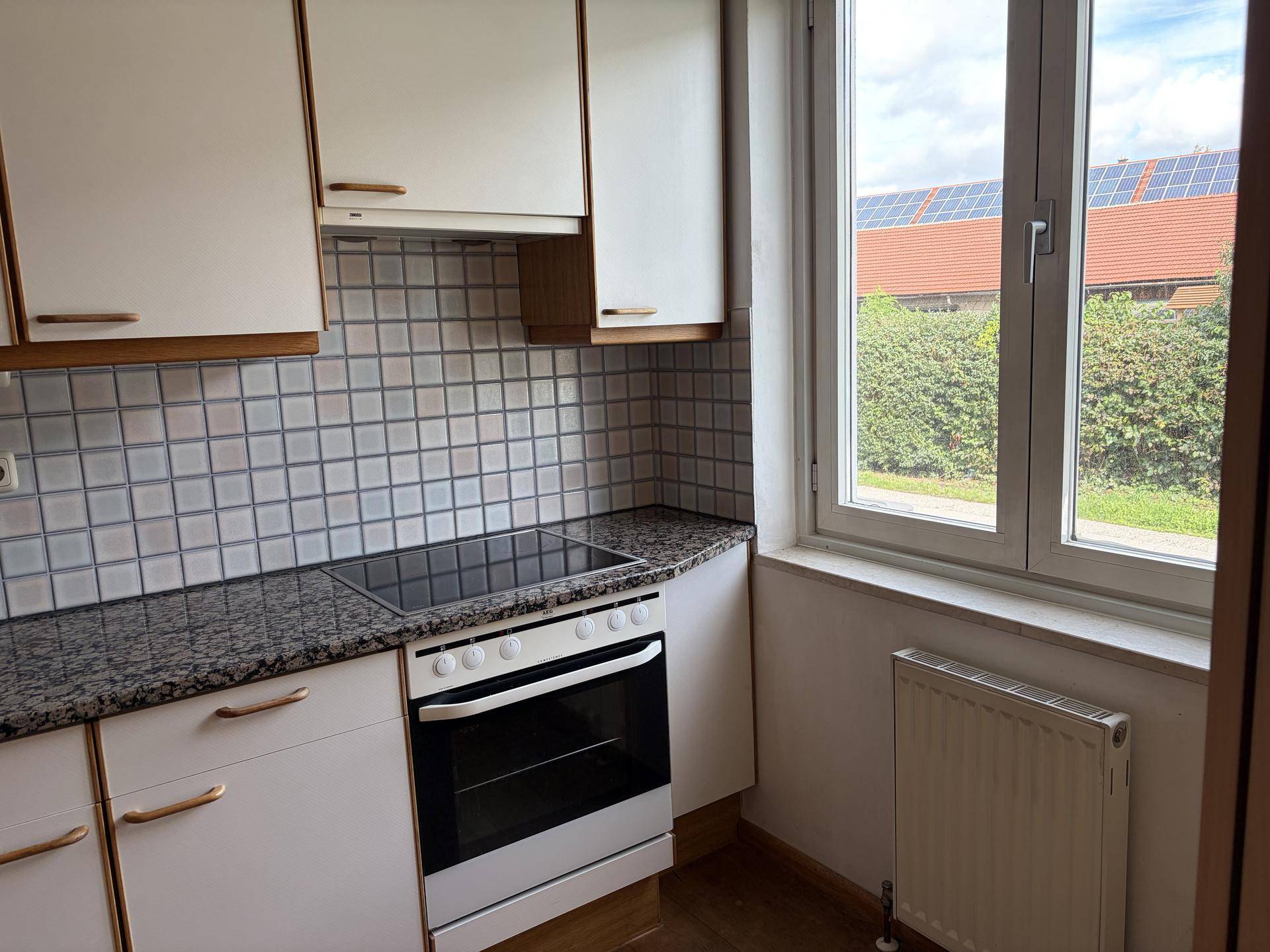 Helle Küche mit weißen Schränken, Kochfeld, Backofen und Fenster mit Blick ins Grüne.