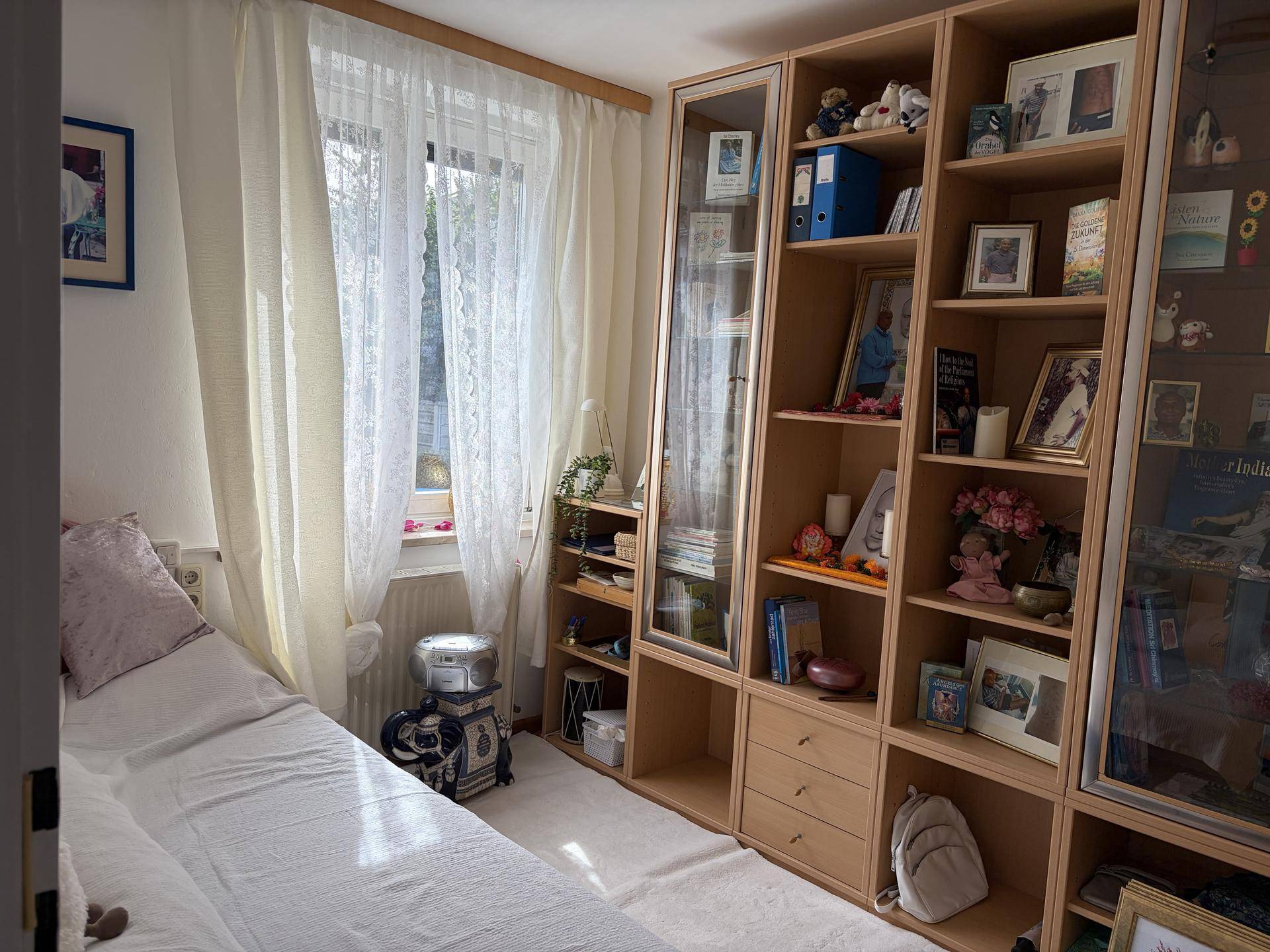 Helles Schlafzimmer mit Fenster, Bücherregal und Teppich auf Holzfußboden.