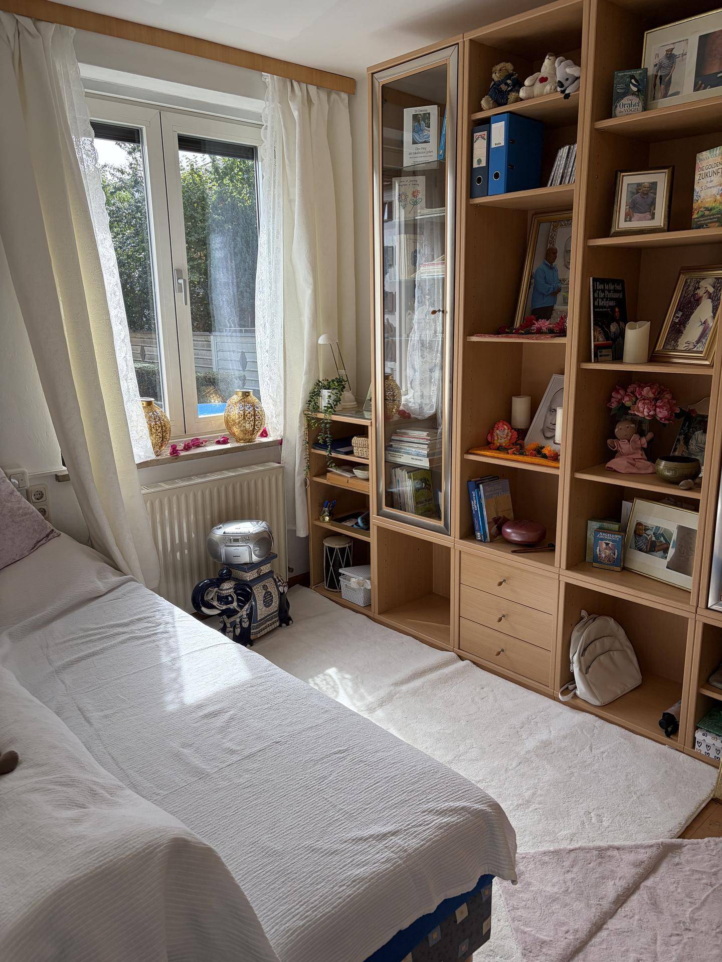 Helles Schlafzimmer mit Fenster, Bücherregal und Teppich auf Holzfußboden.