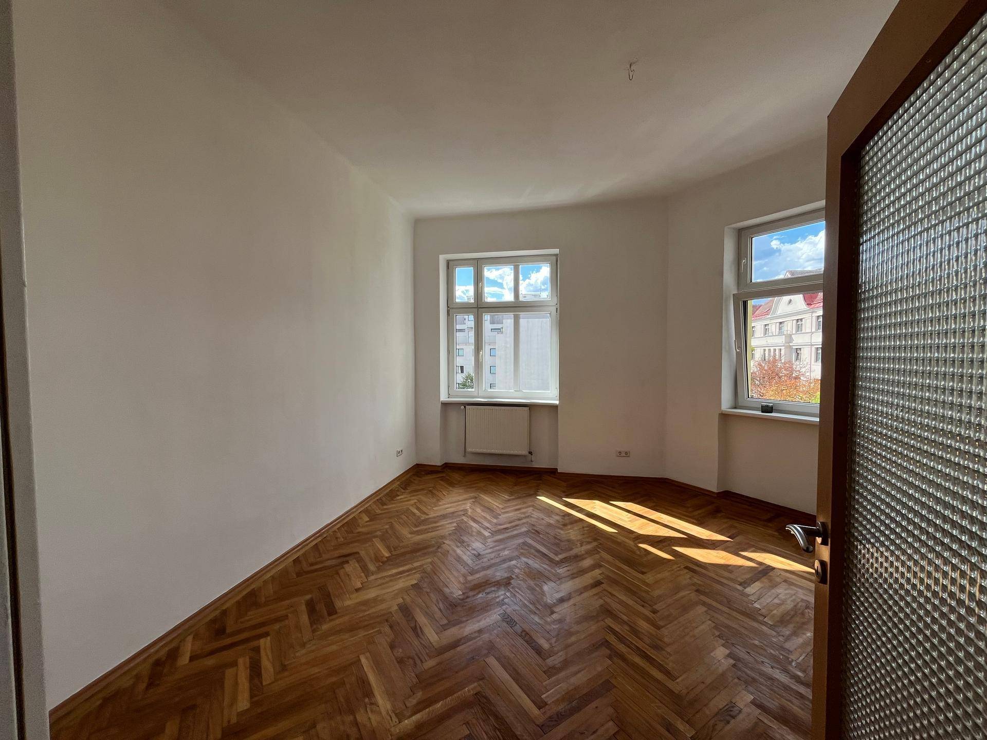 Geräumiges Zimmer mit Fischgrätparkettboden und Blick auf die Straße durch die Fenster.