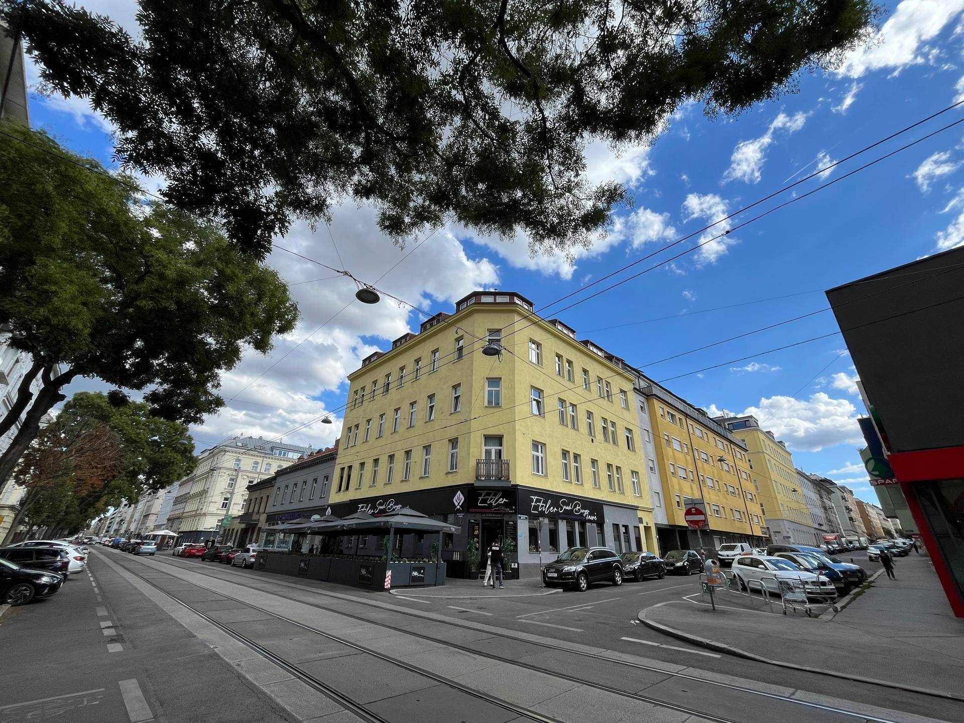 Eckgebäude mit gelber Fassade und Geschäften im Erdgeschoss an einer belebten Straße.