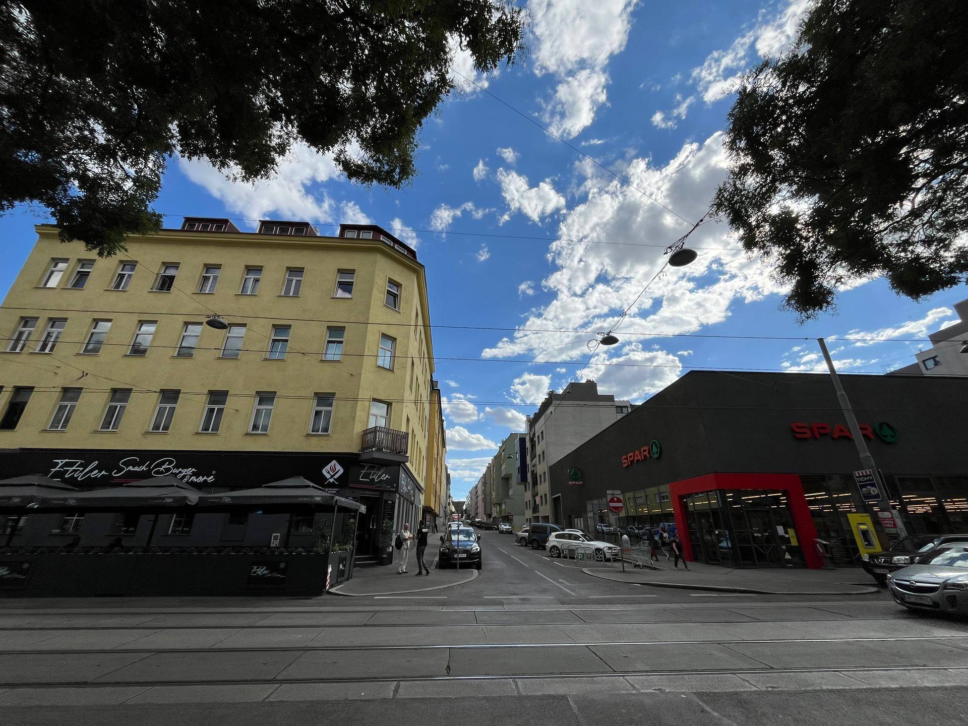 Weitläufige Stadtansicht mit Geschäften, Gebäuden und einem Supermarkt unter blauem Himmel.