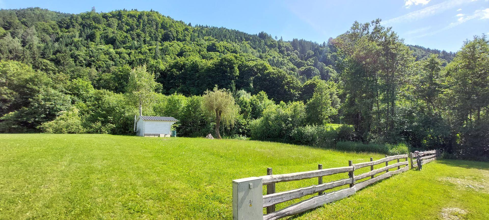 Weitläufige grüne Wiese mit einem kleinen Schuppen am Waldrand und bewaldeten Hügeln im Hintergrund.