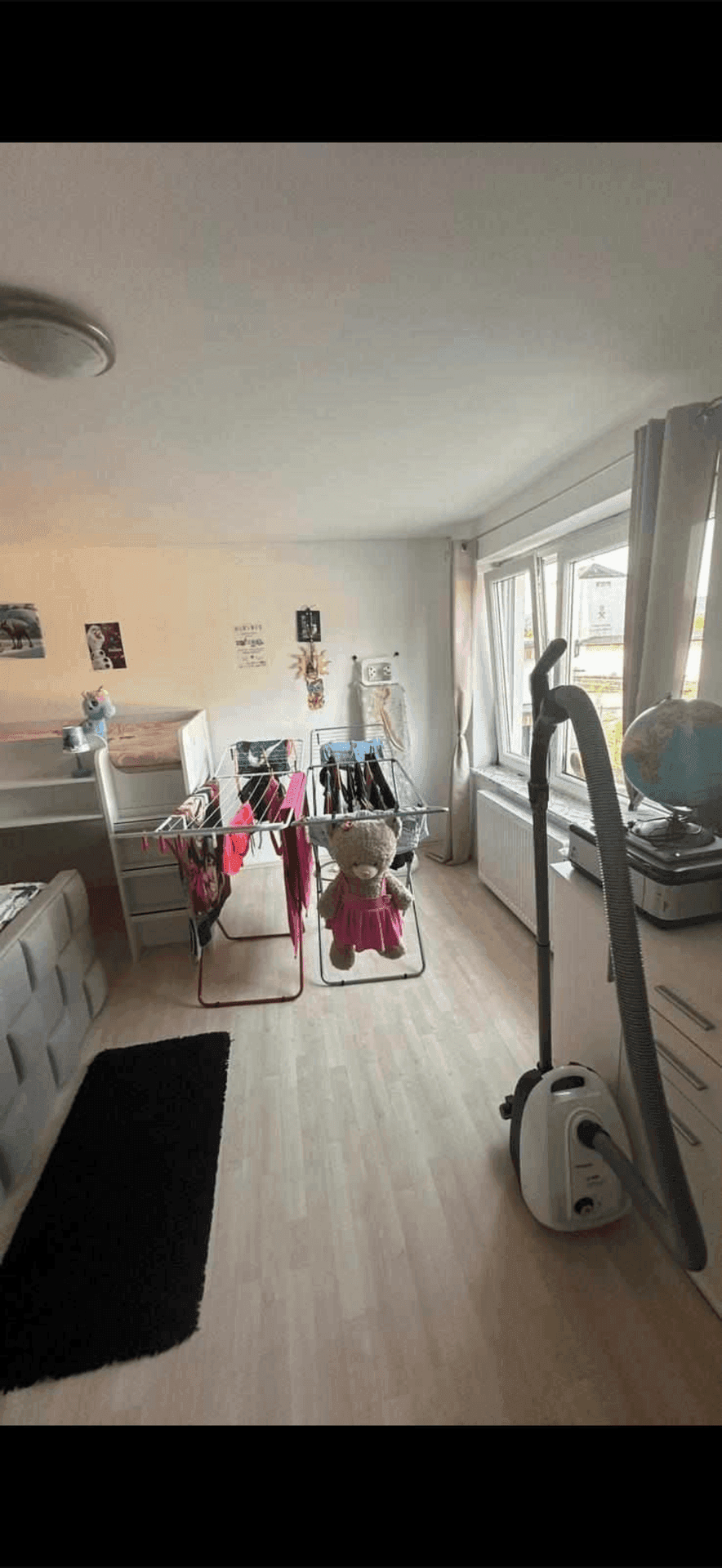 Helles Schlafzimmer mit Wäscheständer, Staubsauger und Fenster mit Blick nach draußen, auf Laminatboden.