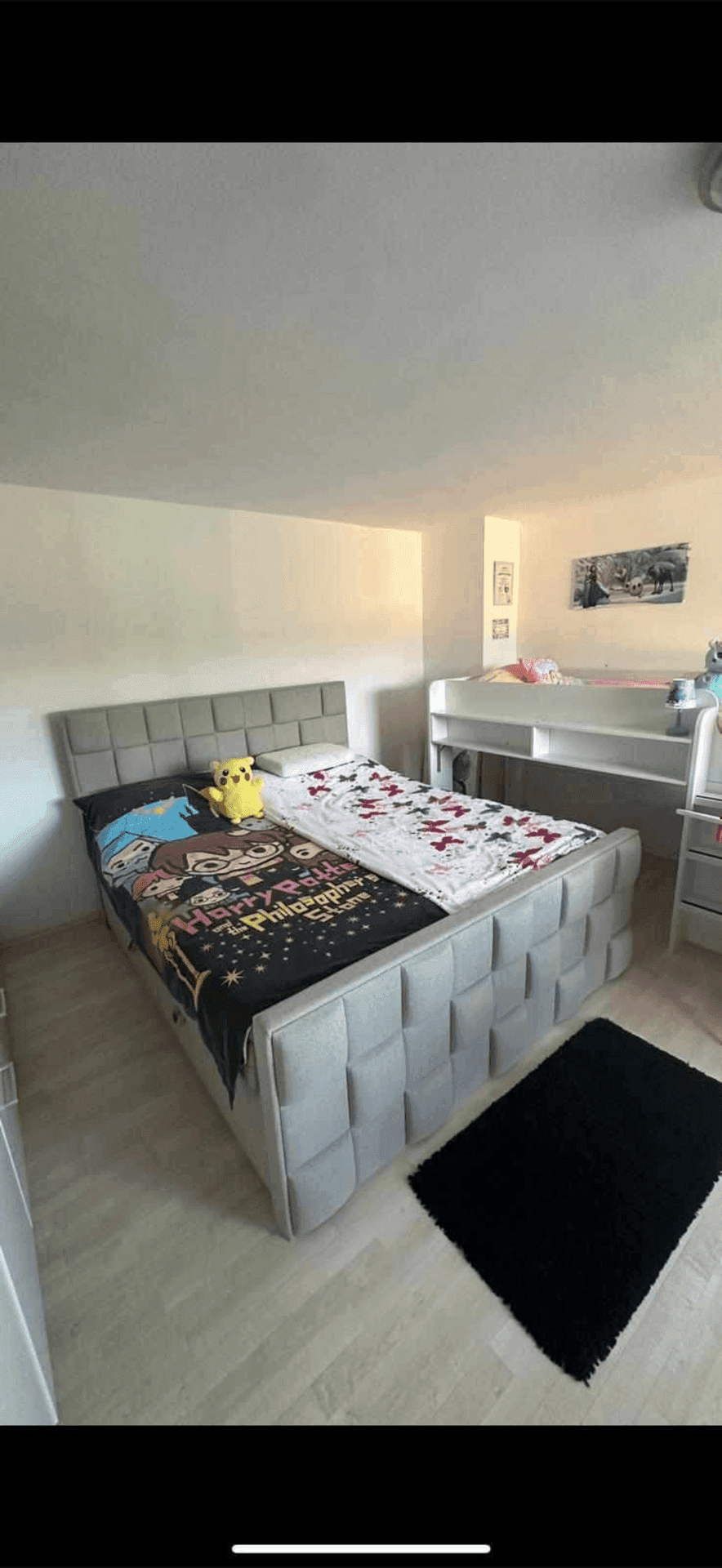 Kinderzimmer mit einem gepolsterten Einzelbett und einem Etagenbett im Hintergrund, auf hellem Laminatboden.