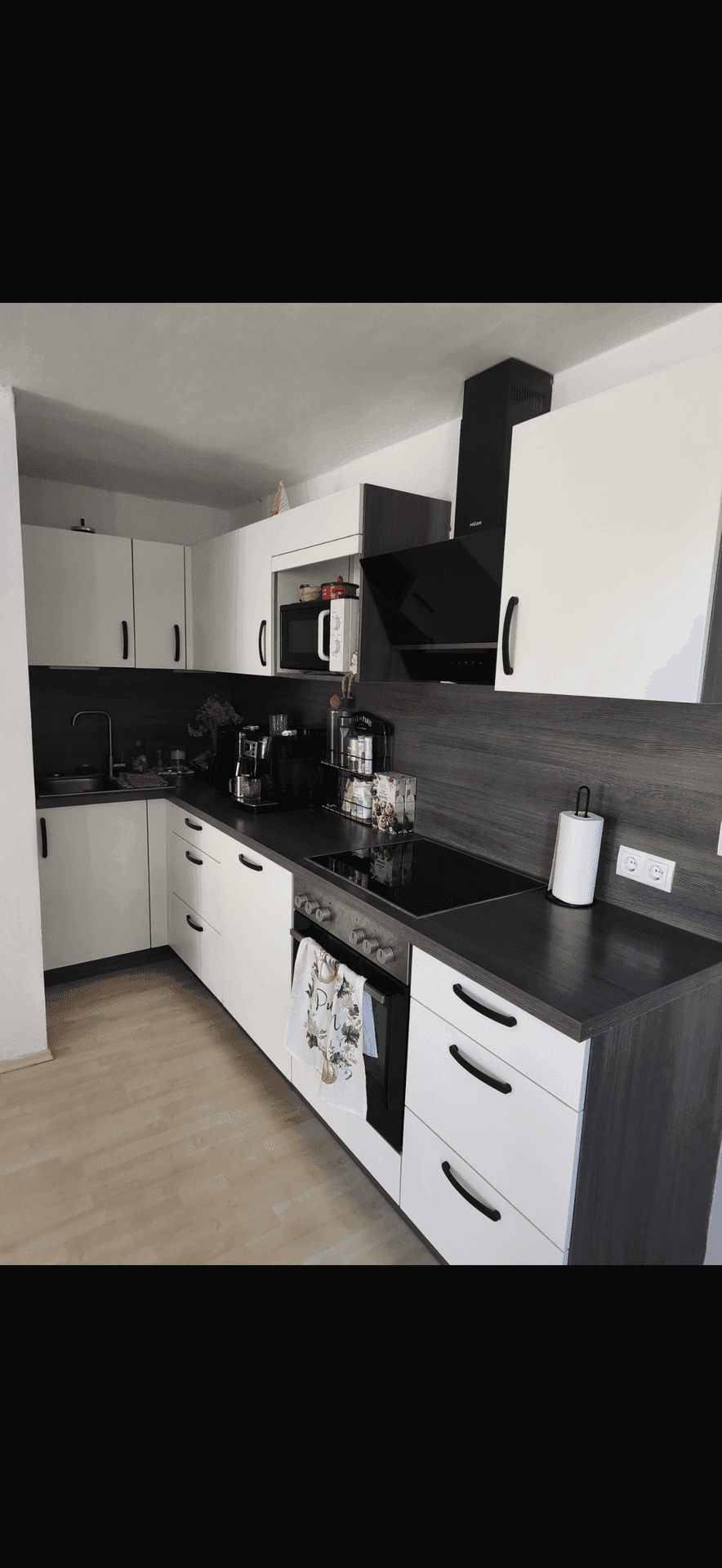 Moderne Küche mit weißen Schränken, dunklen Arbeitsplatten und integrierten Geräten auf hellem Laminatboden.