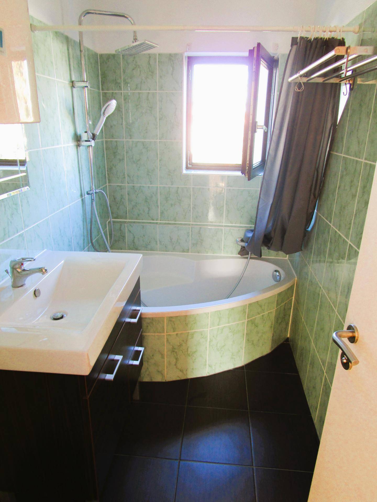 Badezimmer mit grünen Fliesen, Eckbadewanne mit Duschfunktion und einem Fenster.