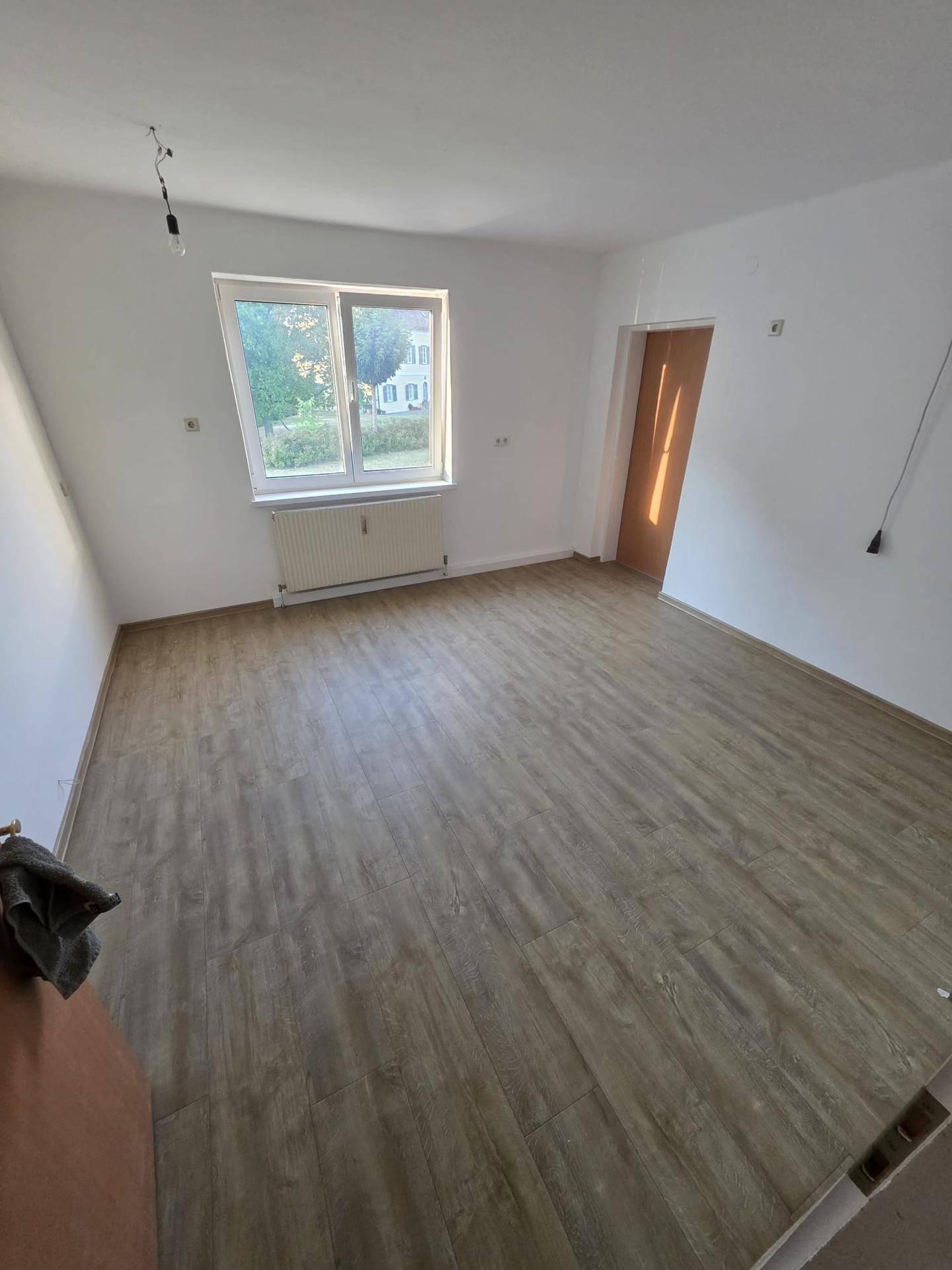 Heller, leerer Raum mit Laminatboden und einem Fenster, das Tageslicht hereinlässt.