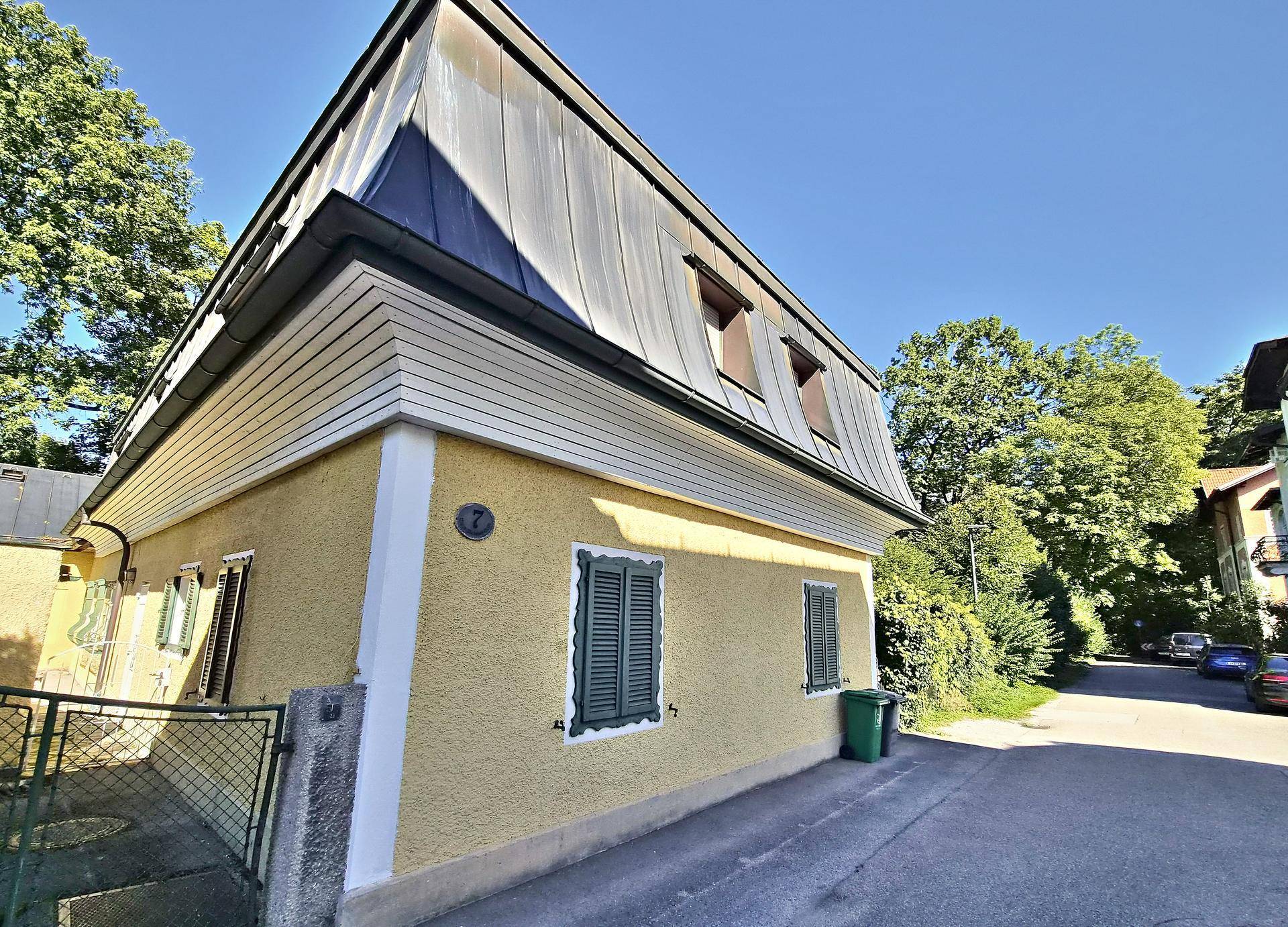 Seitenansicht eines gelben Hauses mit grünen Fensterläden und einem markanten Mansarddach.