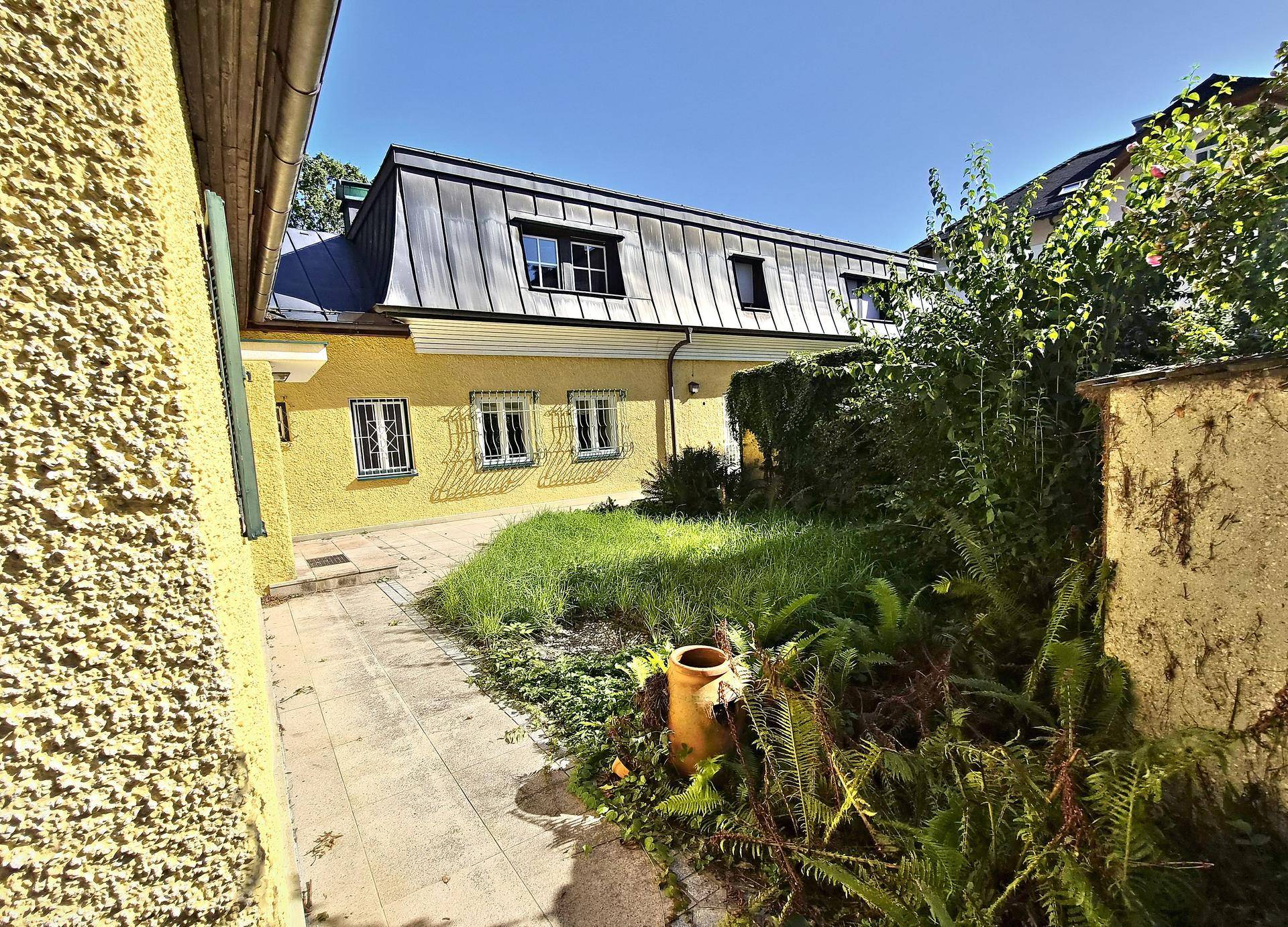 Blick auf den hinteren Teil des Hauses mit Mansarddach und einem verwilderten Gartenbereich mit gepflastertem Weg.
