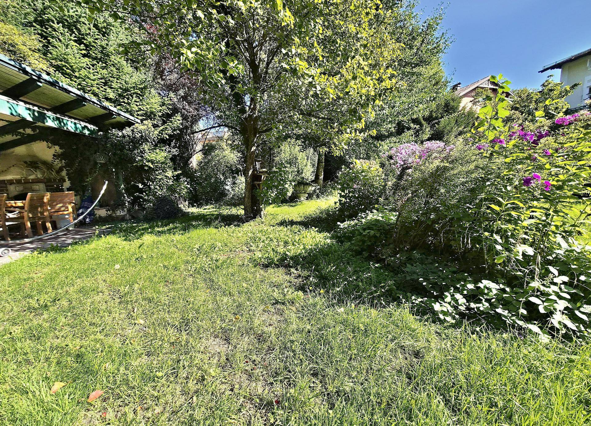 Weitläufiger, sonniger Garten mit altem Baum, blühenden Sträuchern und einem überdachten Sitzbereich.