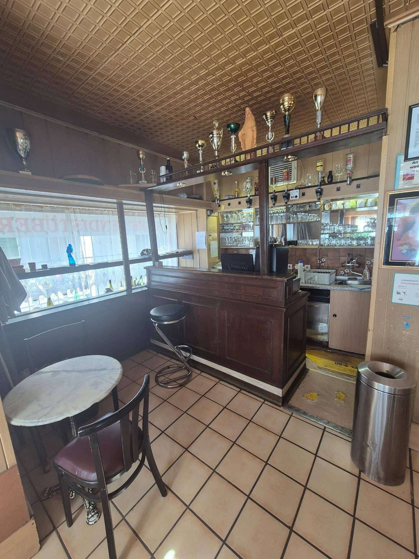 Klassische Bar mit Holztheke, Hockern und Regalen voller Gläser und Flaschen, mit Fliesenboden.