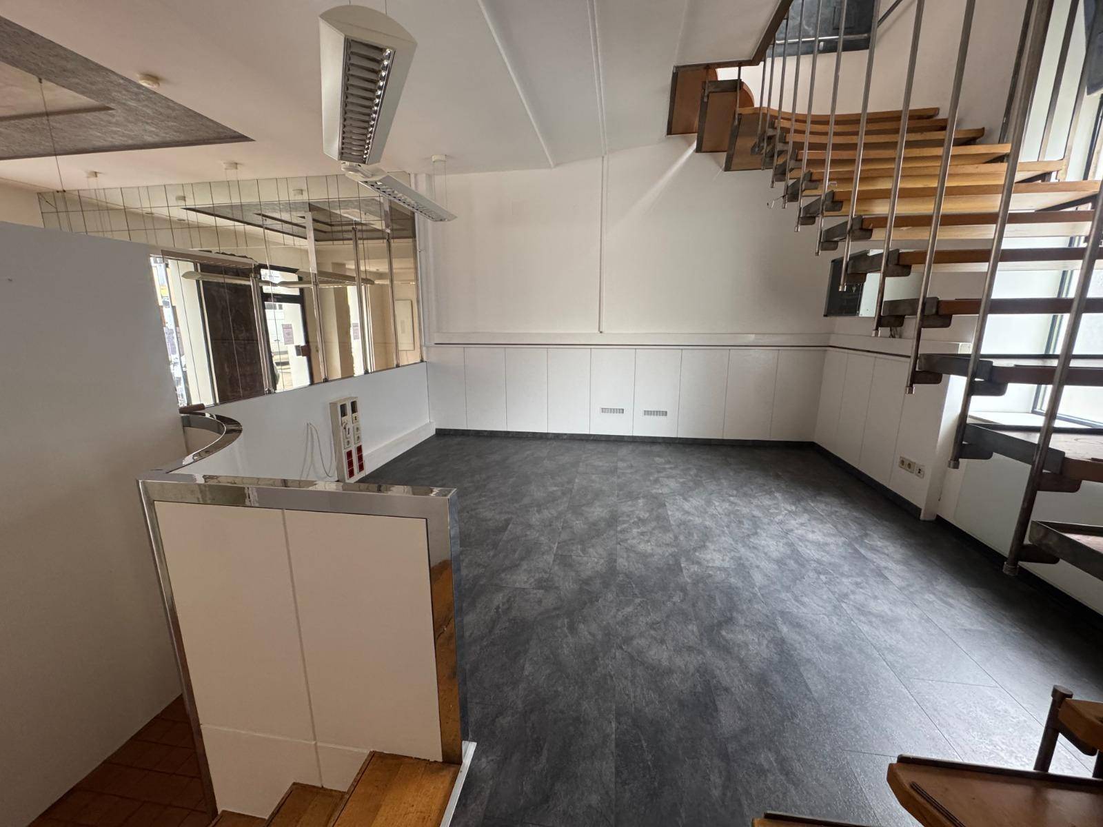 Geräumiger Gewerberaum mit dunklem Vinylboden und einer modernen Treppe zum Obergeschoss.
