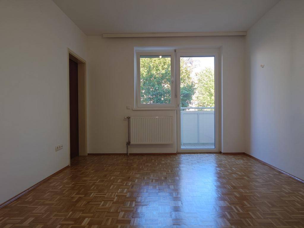 Geräumiges Wohnzimmer mit Parkettboden, weißen Wänden und Zugang zu einem Balkon.