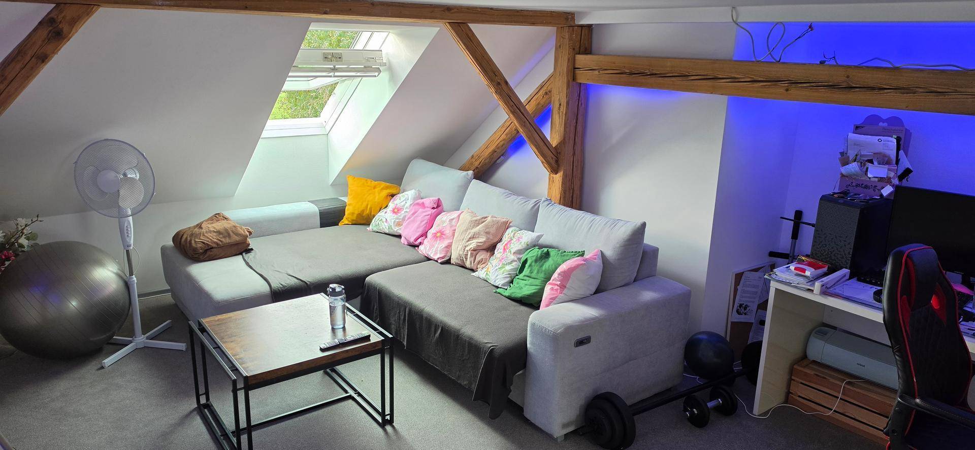 Gemütliches Wohnzimmer im Dachgeschoss mit Sofa, Schreibtisch und sichtbaren Holzbalken.