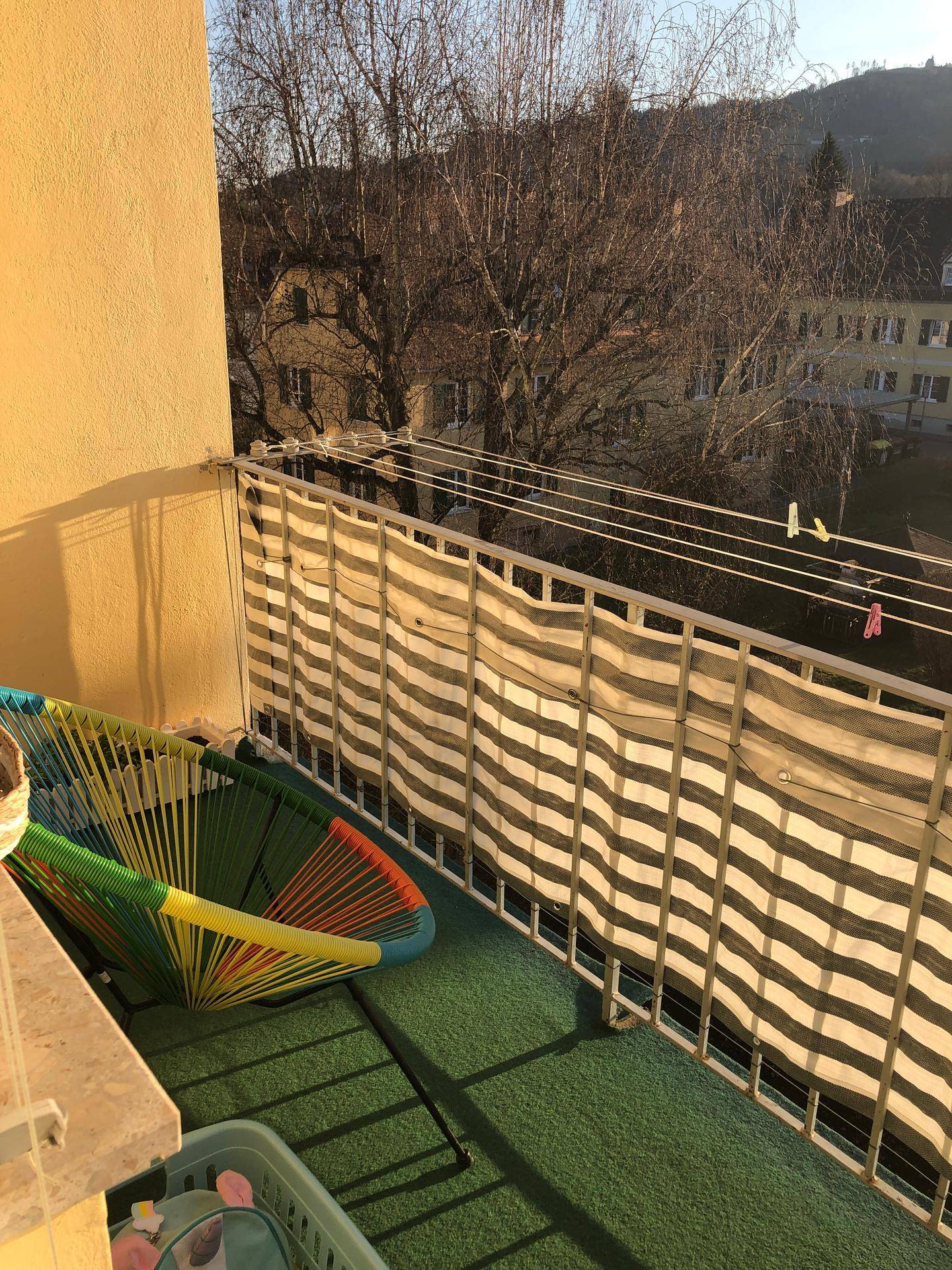 Sonniger Balkon mit Kunstrasen, bunter Sitzgelegenheit und Blick ins Grüne.