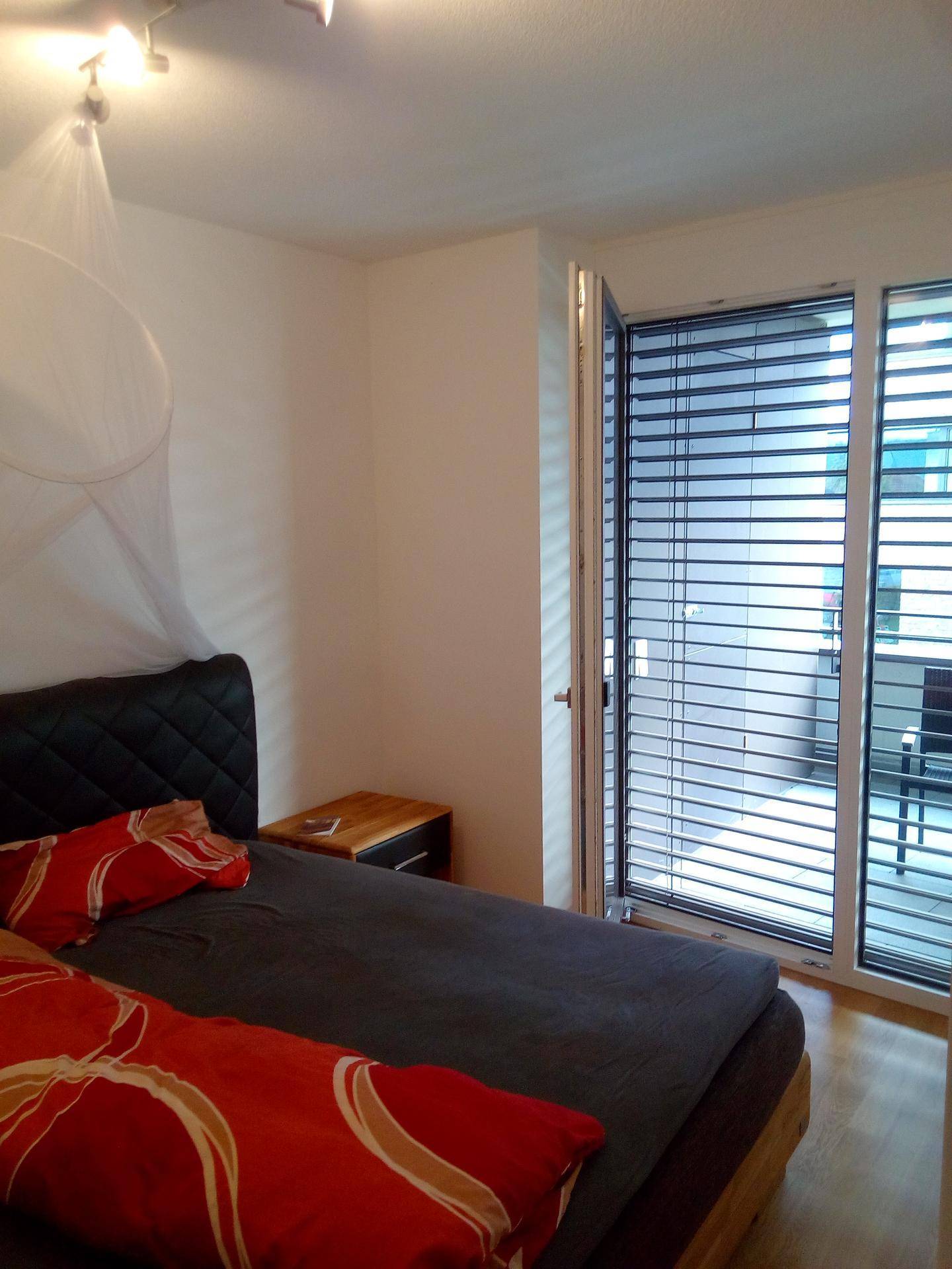 Schlafzimmer mit Doppelbett, Nachttisch und großen Fenstern mit Jalousien für Privatsphäre.
