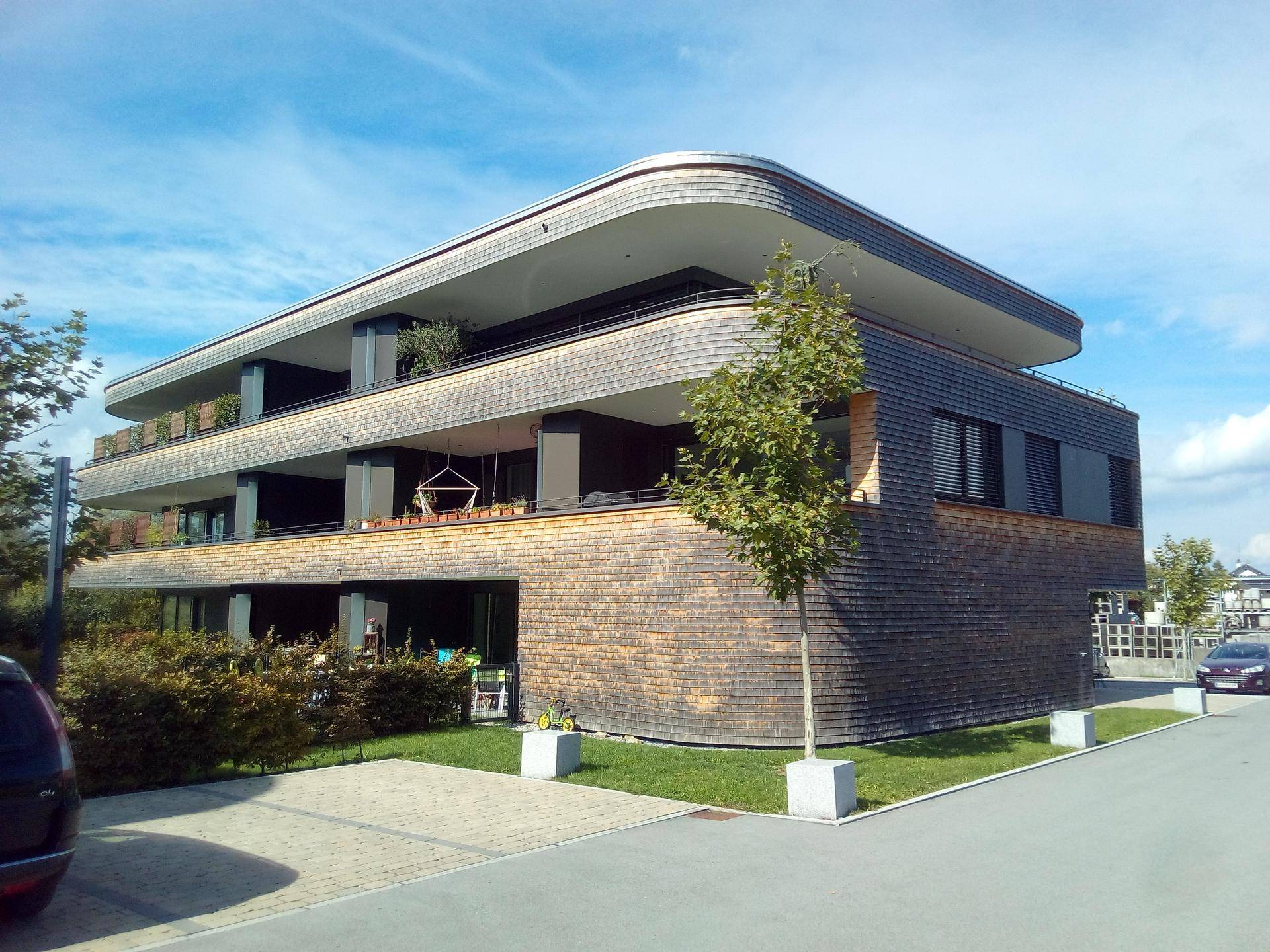 Modernes Mehrfamilienhaus mit geschwungenem Dach und Holzschindelfassade in grüner Umgebung.