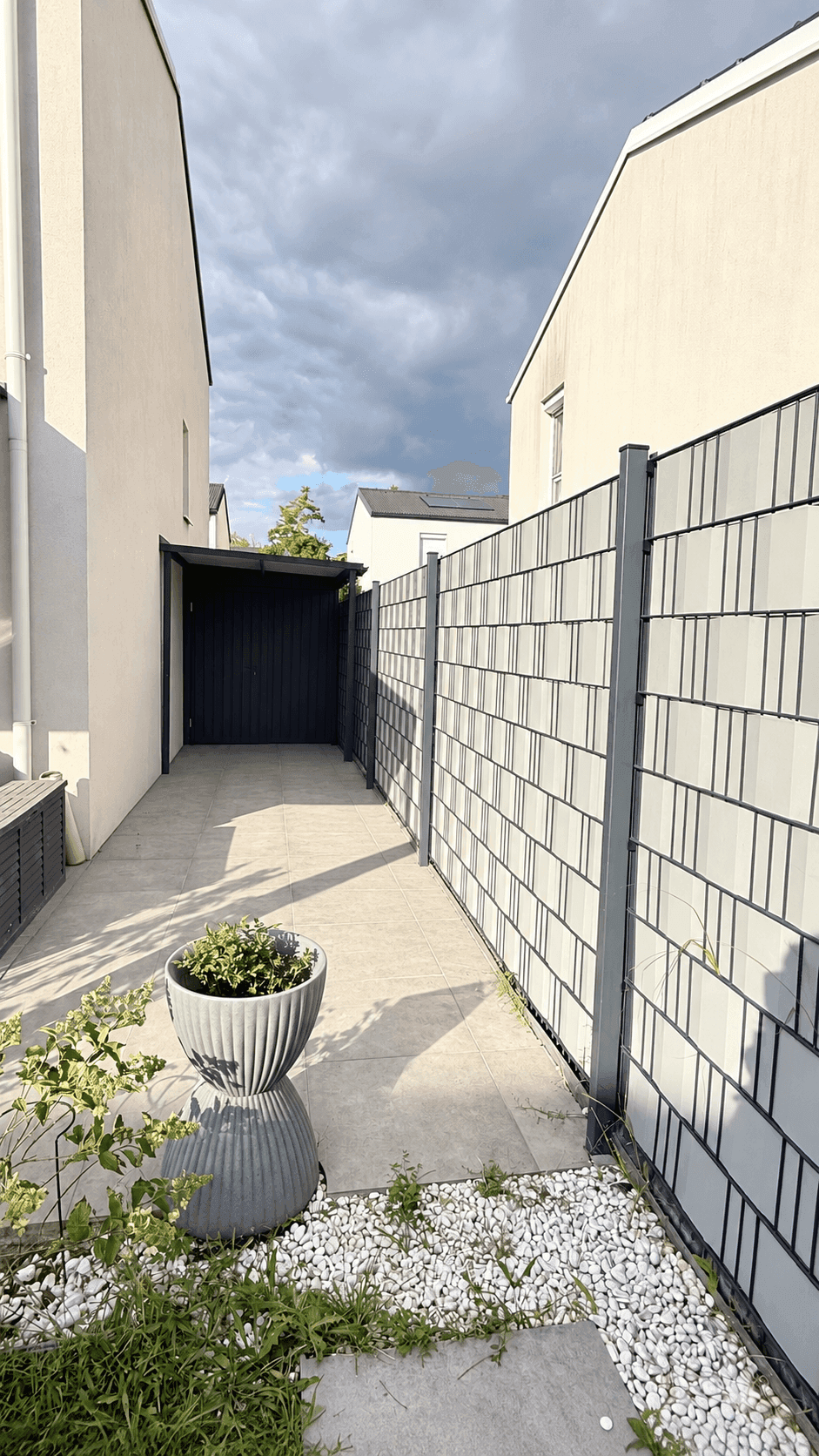 Seitlicher Zugangsweg zum Haus mit modernem Zaun und einem Carport im Hintergrund.